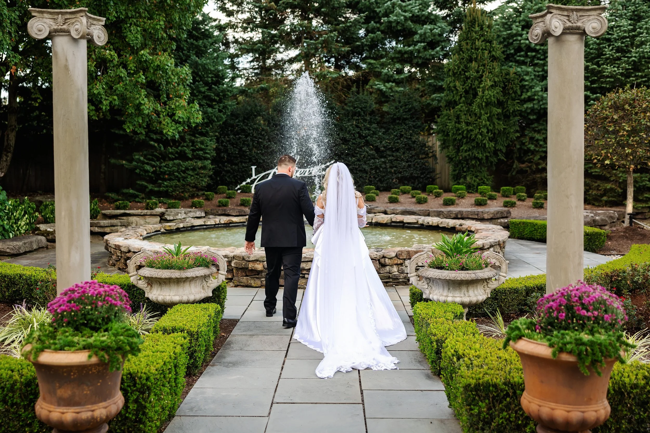 A newlywed couple walking hand in hand towards a fountain in a landscaped garden, with the bride in a white wedding dress and the groom in a dark suit, surrounded by potted flowers and greenery.