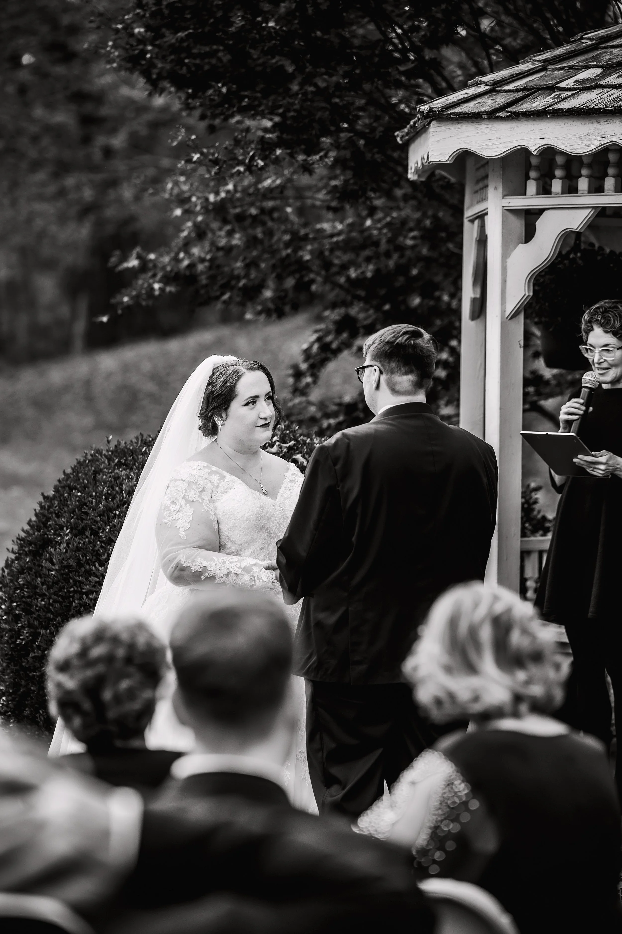 A black and white photo of a wedding ceremony outdoors with the bride and groom standing and exchanging vows. The bride is in a lace wedding dress and veil, and the groom in a dark suit. An officiant is speaking into a microphone nearby, and guests are seated watching the ceremony.