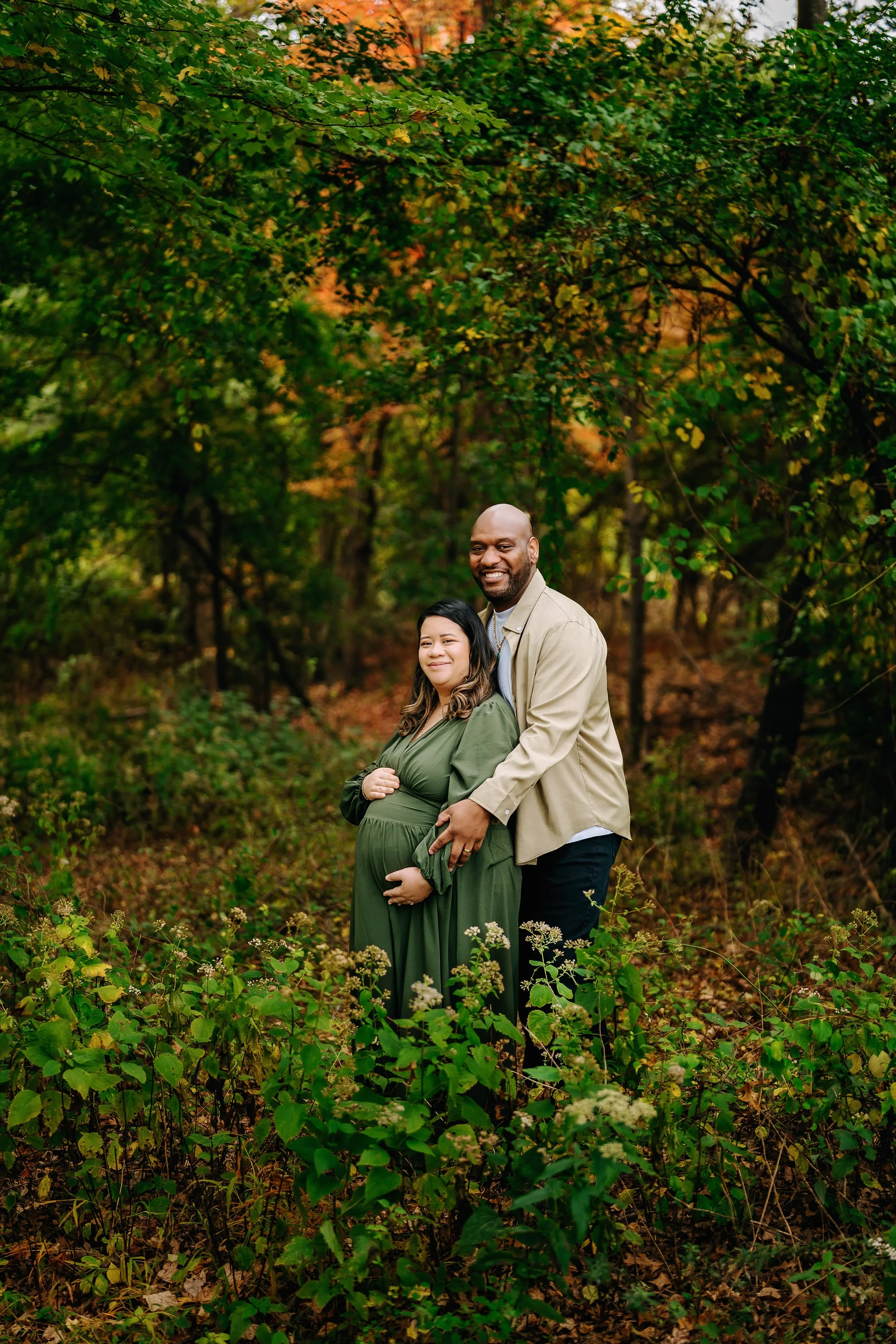 A smiling pregnant woman in a green dress standing in a forest with a man behind her, both enjoying autumn foliage.