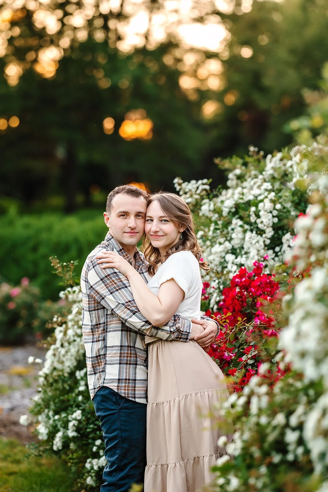 A young couple embraces outdoors with blooming flowers and trees at sunset in the background.