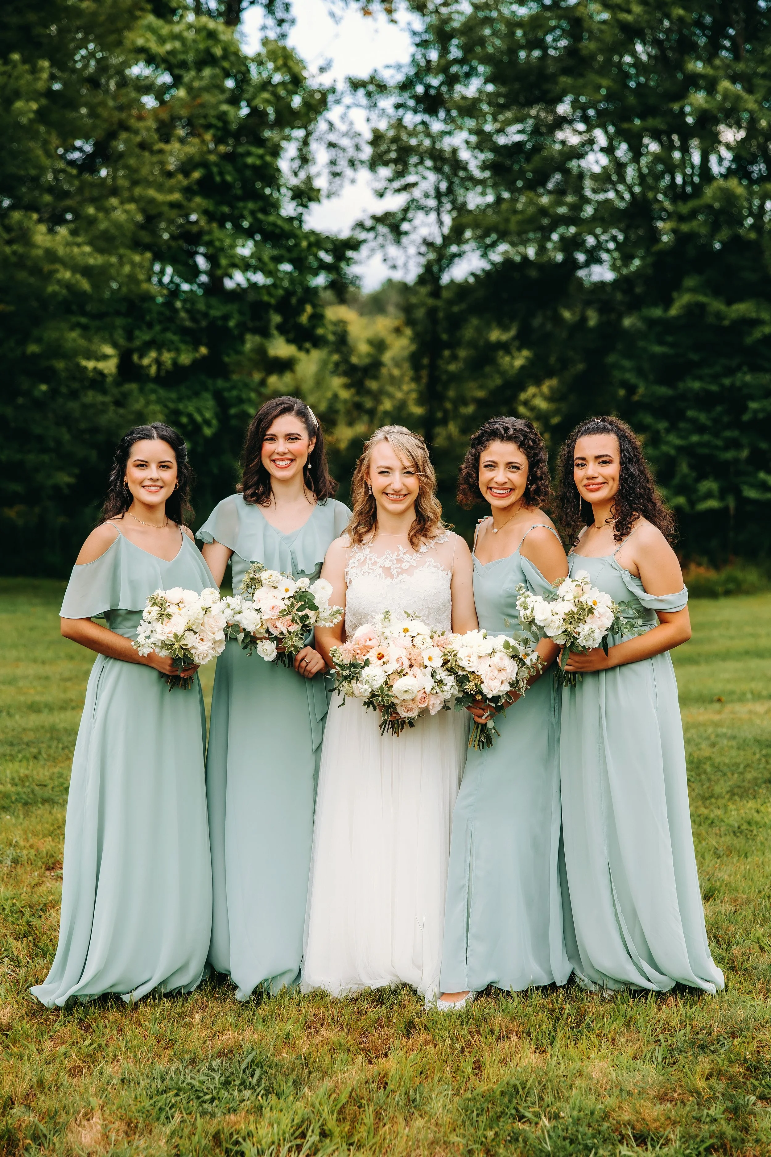A bride in a white wedding dress standing with five bridesmaids in matching light blue dresses, all holding bouquets of flowers, outdoors on a grassy area with trees in the background.
