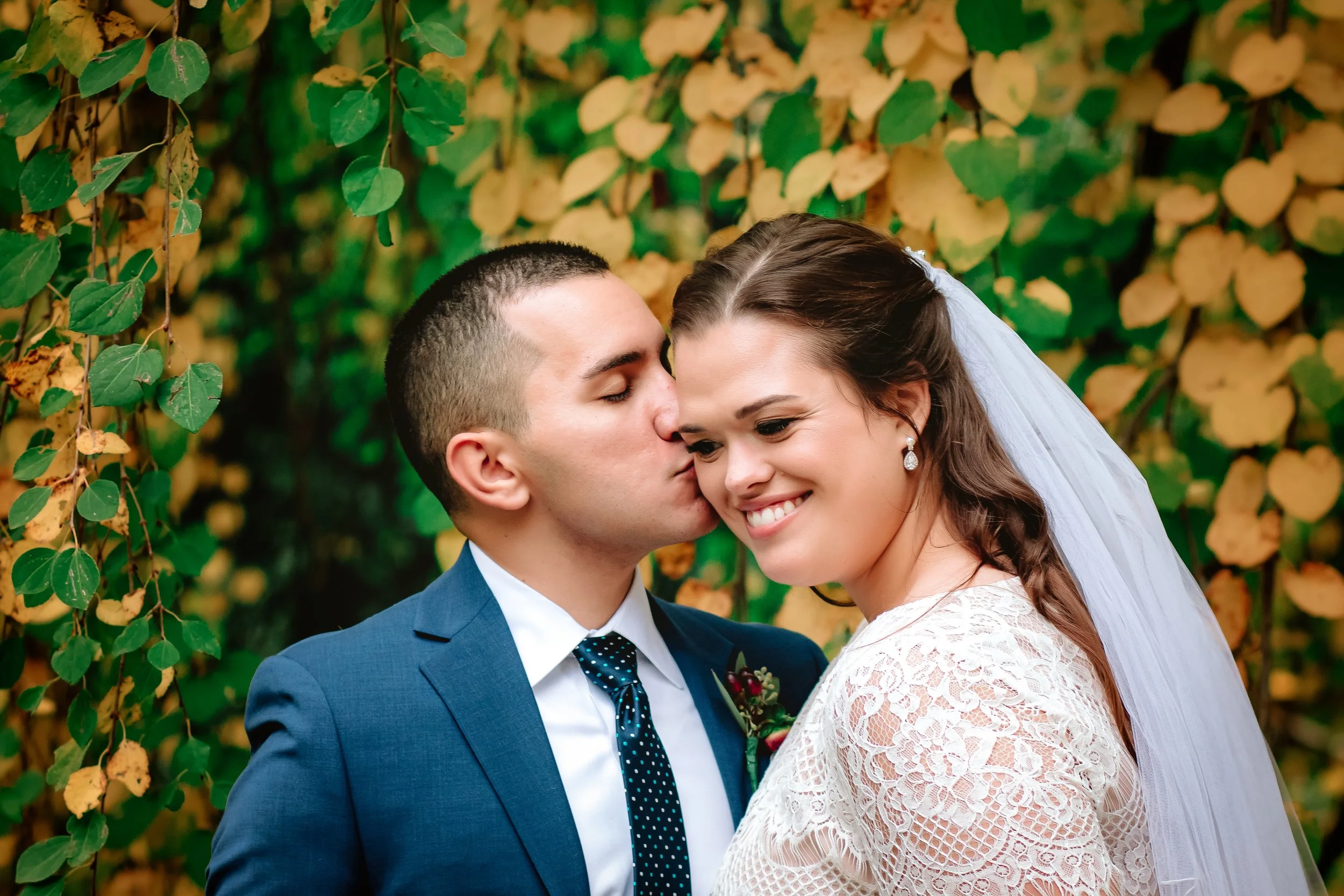 A bride and groom on their wedding day, with the groom kissing the bride on the cheek. The bride is smiling, wearing earrings and a veil, and the groom is in a blue suit with a tie, standing in front of a leafy, green and yellow background.