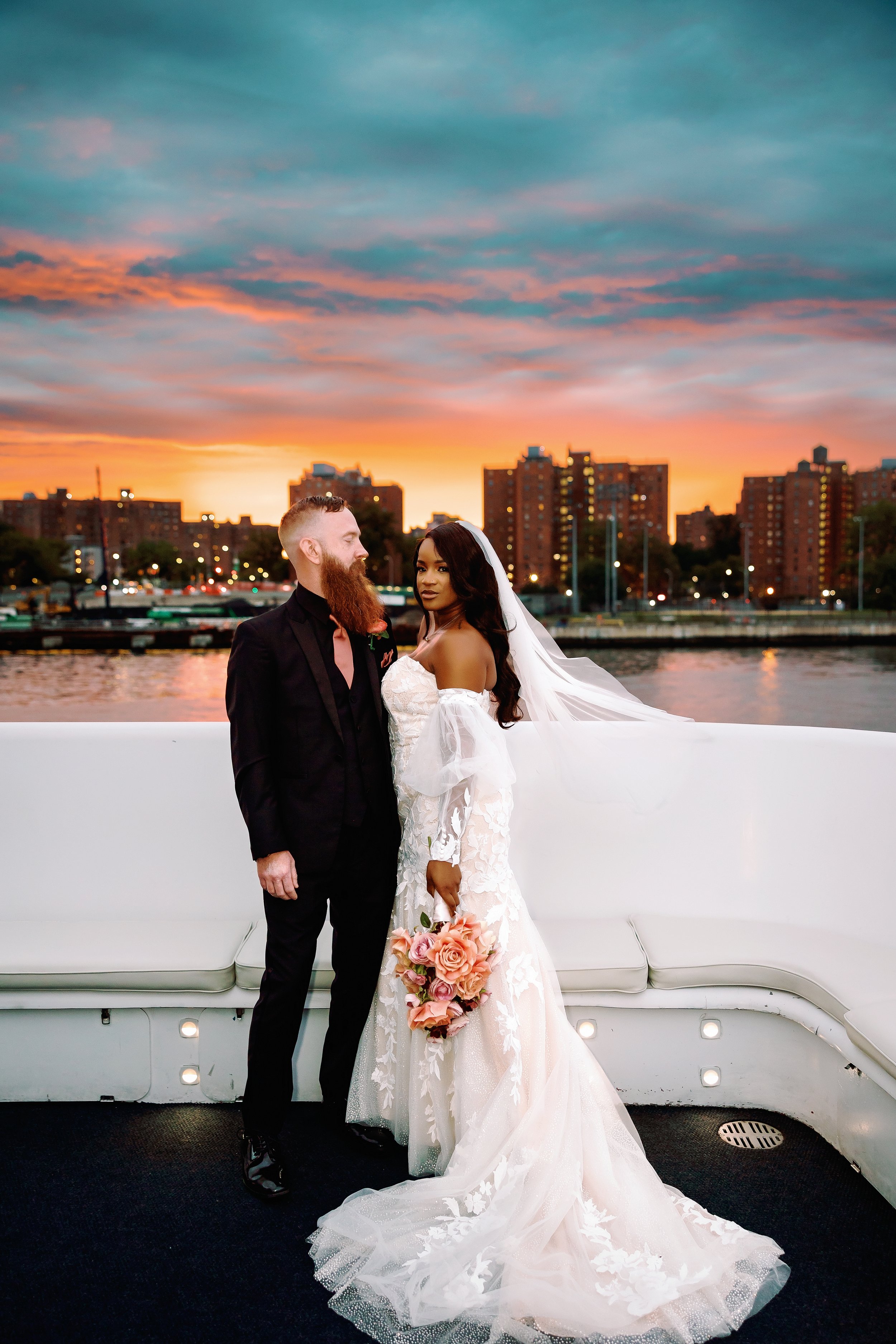 Bride and groom on a boat at sunset, with city buildings and water in the background.