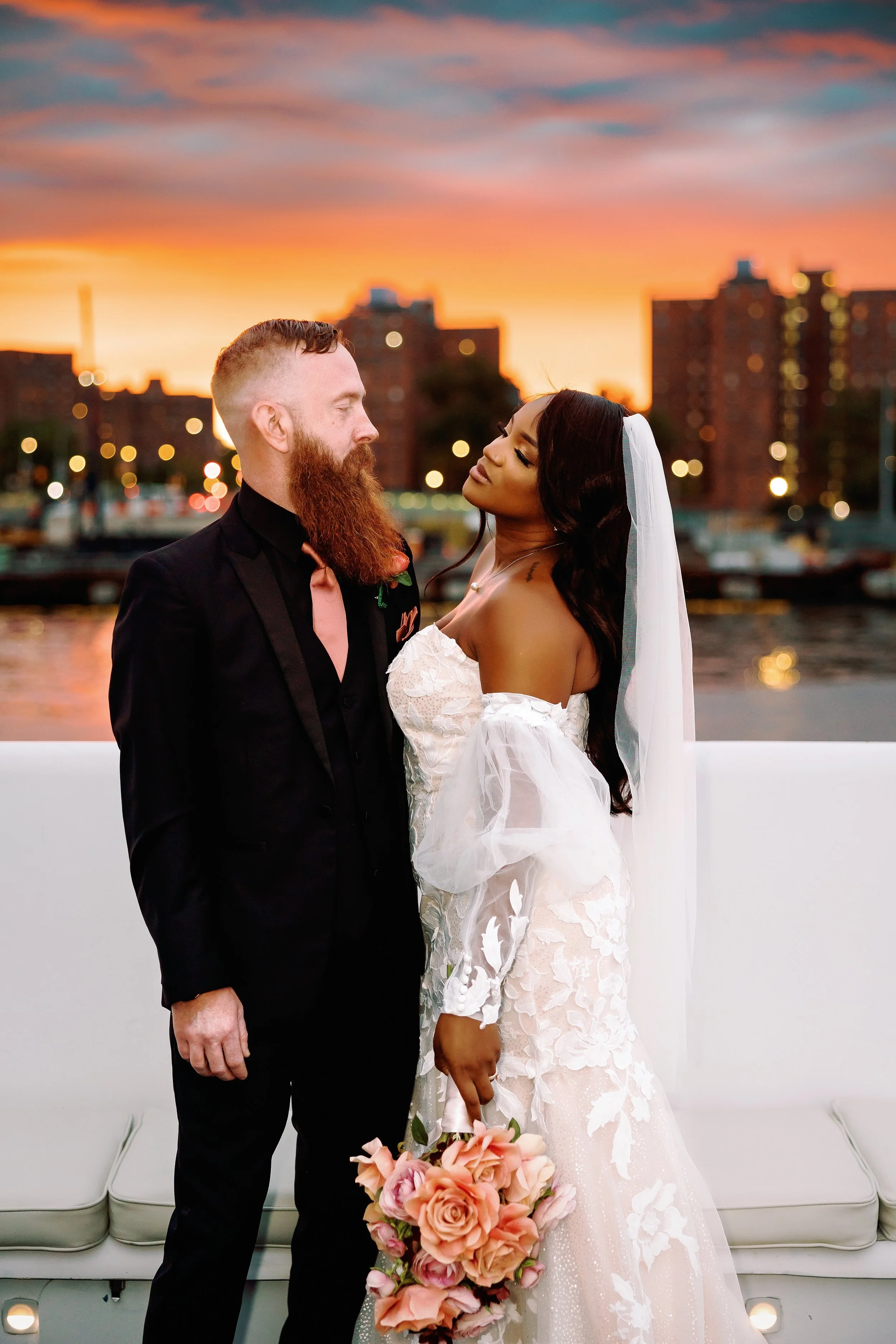 A bride and groom stand close together during sunset by the water, with city buildings and a colorful sky in the background. The bride wears a lace wedding gown and veil, holding a bouquet of pink roses, and looks at the groom. The groom wears a black suit with a pink tie and has a red beard.