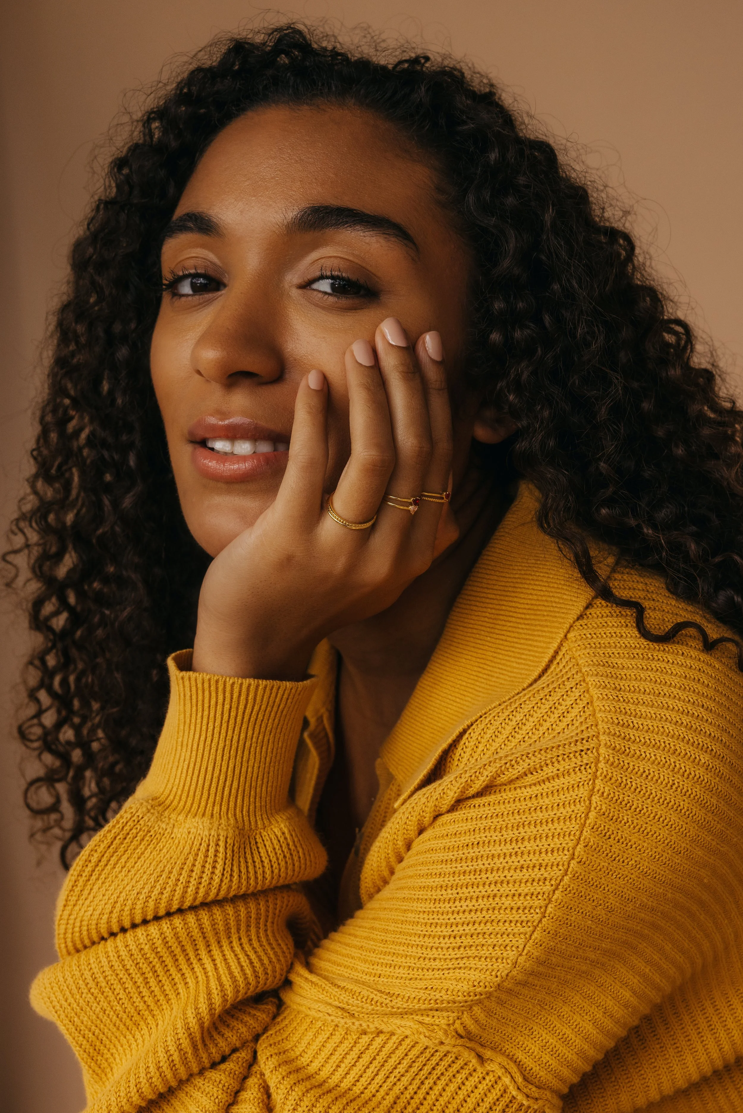 Close-up of a person with curly hair wearing a yellow sweater, resting their chin on their hand with rings on their fingers.