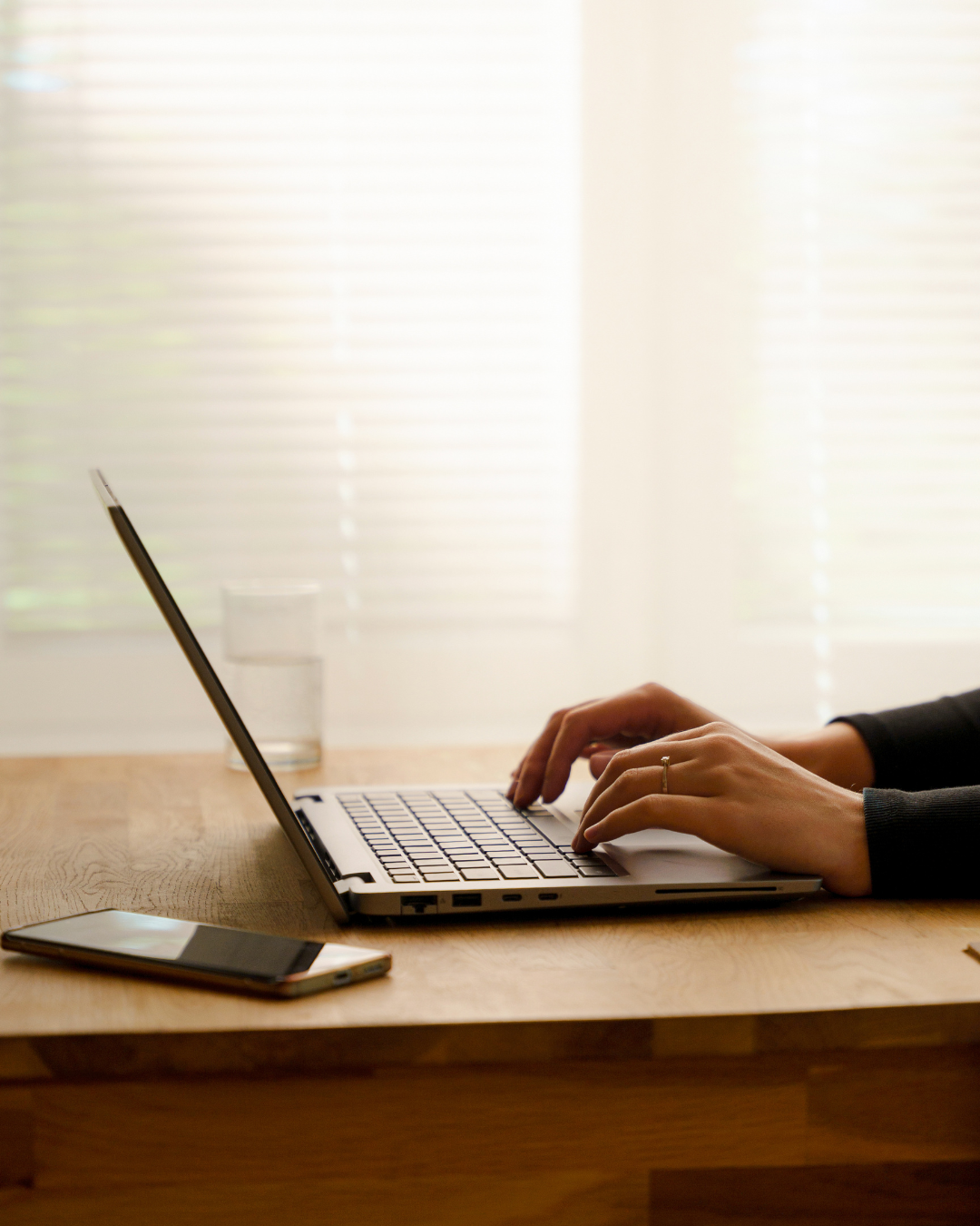 Person typing on a laptop at a wooden desk, with a smartphone nearby and a glass of water in the background.