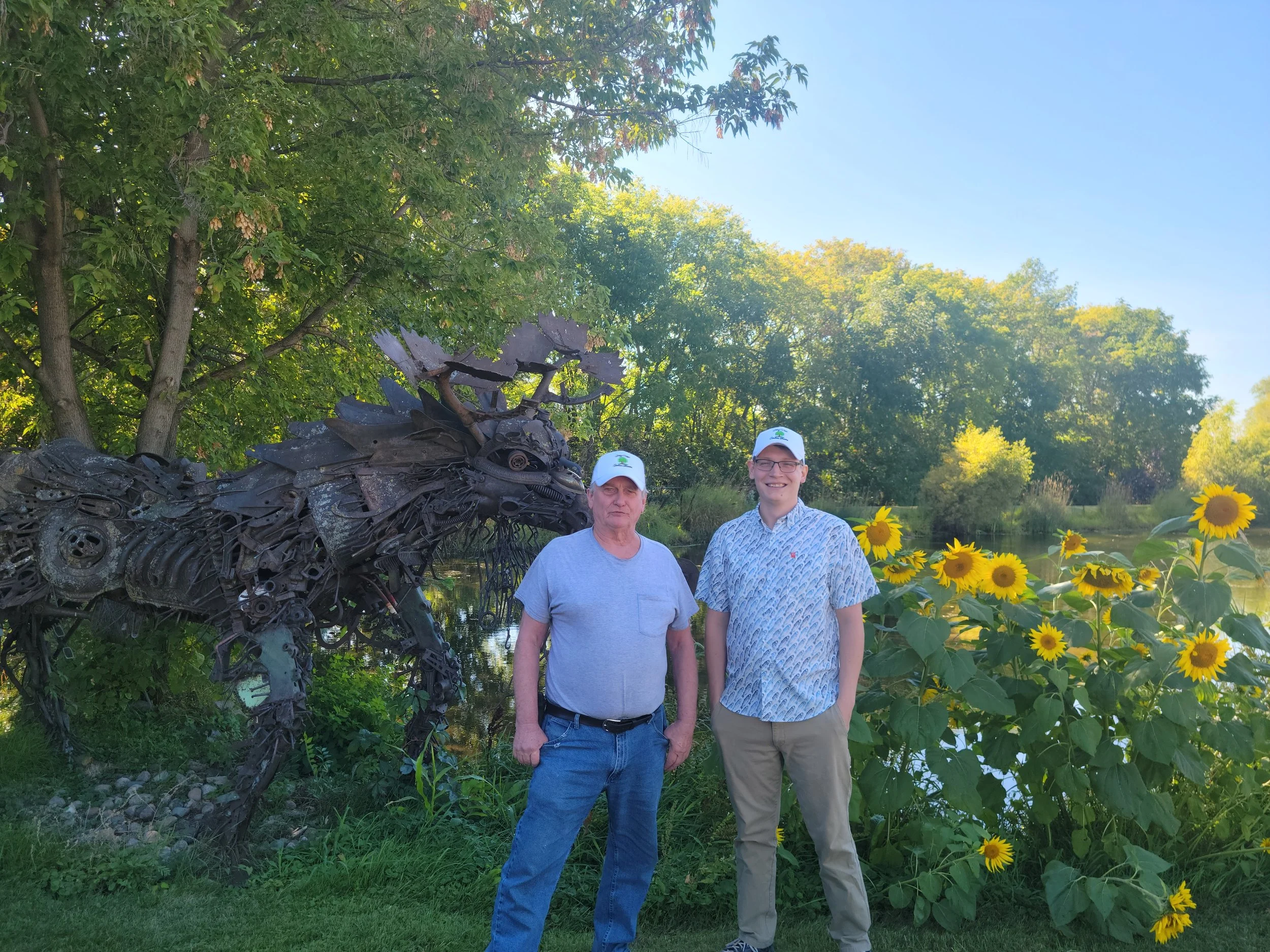 Two men posing outdoors beside a sculpture of a moose made from scrap metal, with sunflowers and trees in the background.