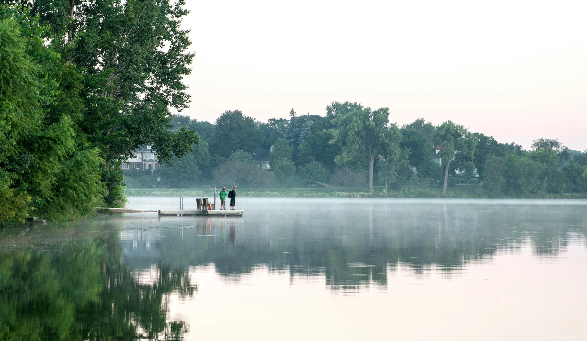 Two people fishing on a mist-covered lake in a serene landscape, with trees and houses in the background.