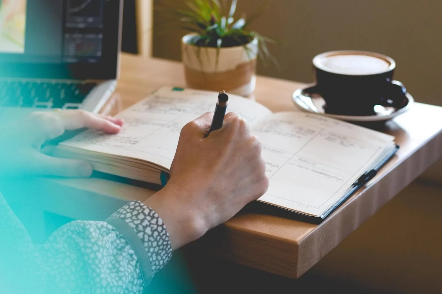 Photo of woman's hand writing in a planner next to a laptop and cup of coffee