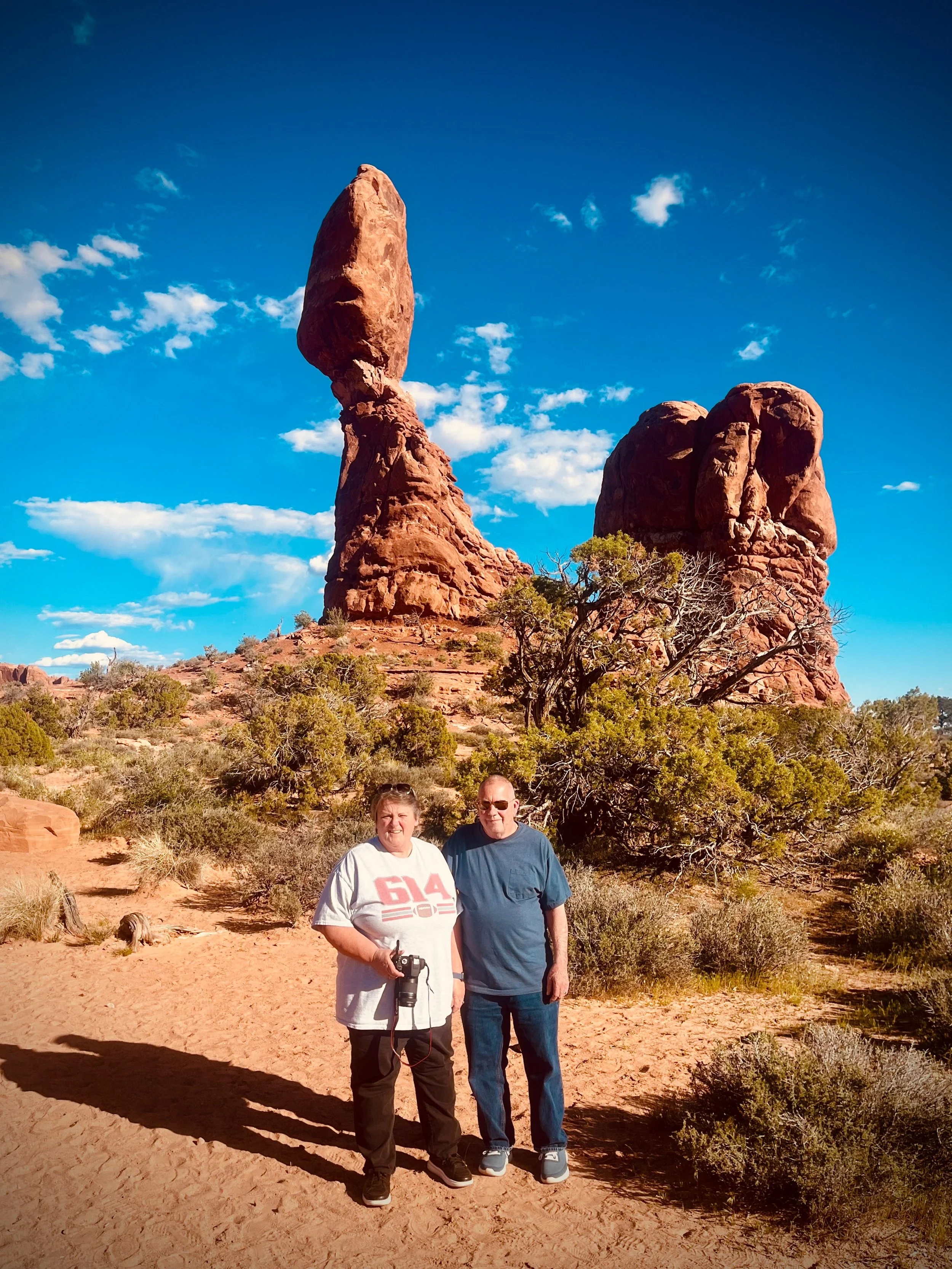 Two people standing on a dirt trail in front of balanced rock formations in a desert landscape with clear blue sky and scattered clouds.