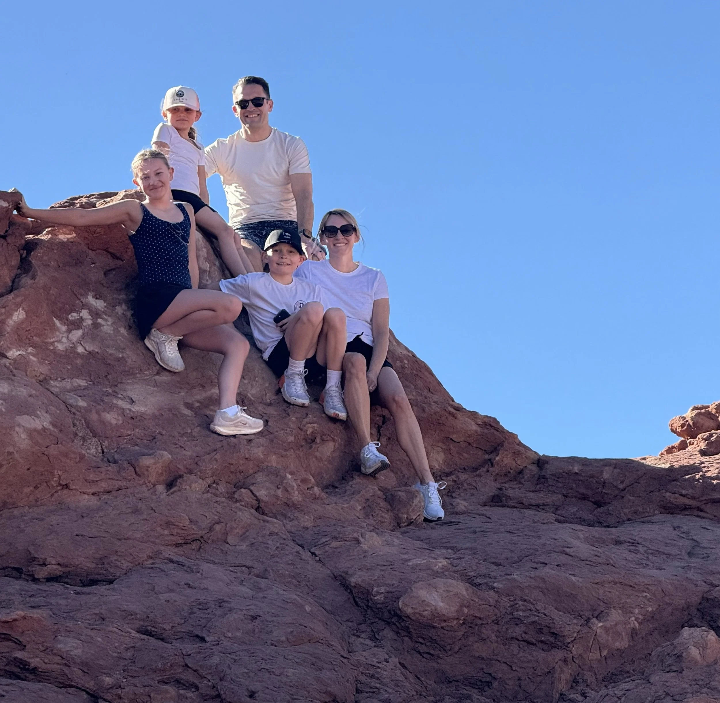 A family of six sitting and standing on red rock formations outdoors under a clear blue sky, smiling at the camera.