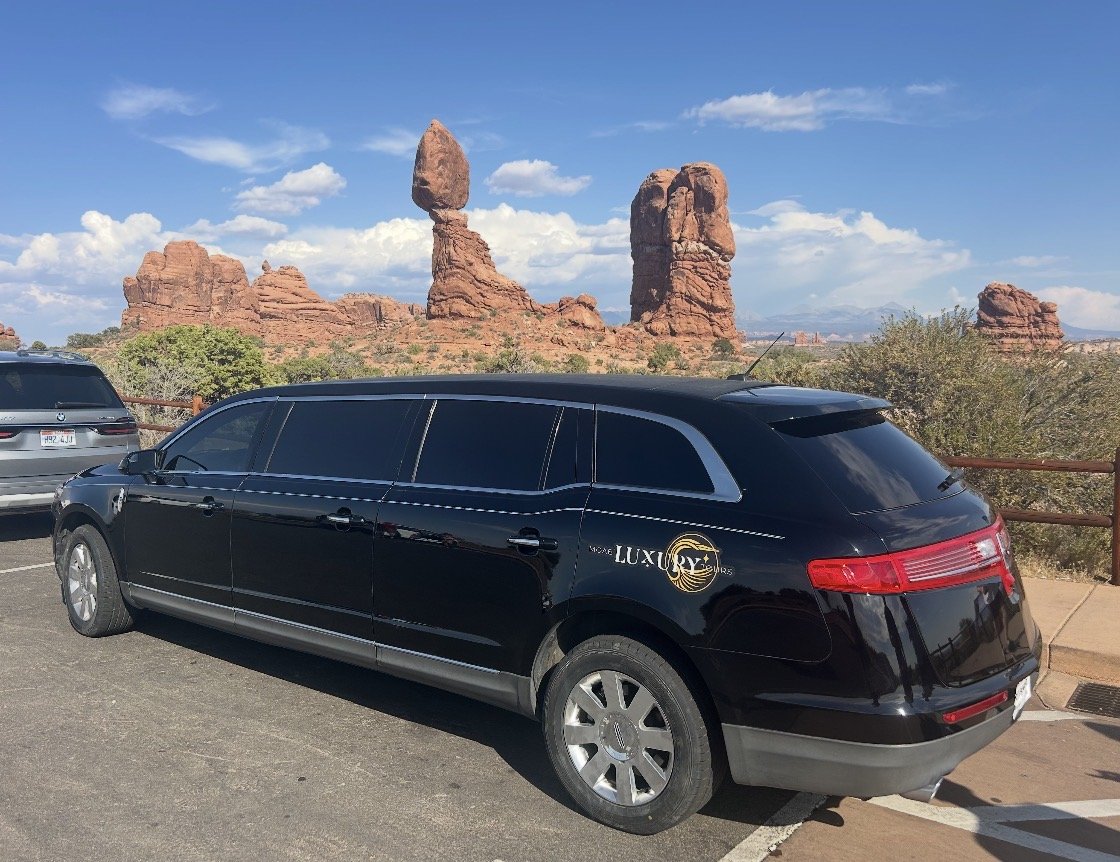 A black limousine with a luxury logo parked in front of the red rock formations of Arches National Park in Utah.