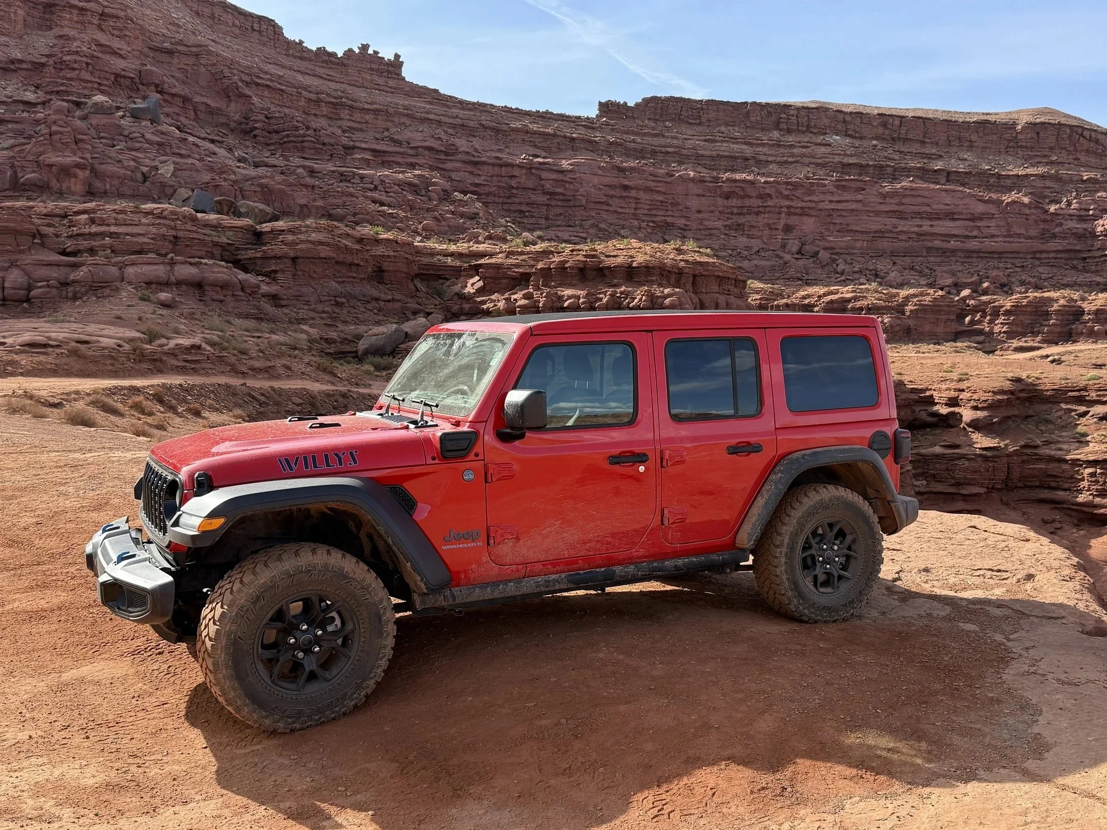 Red Jeep Wrangler with muddy tires parked on a dirt path in a desert landscape with red rock formations in the background.