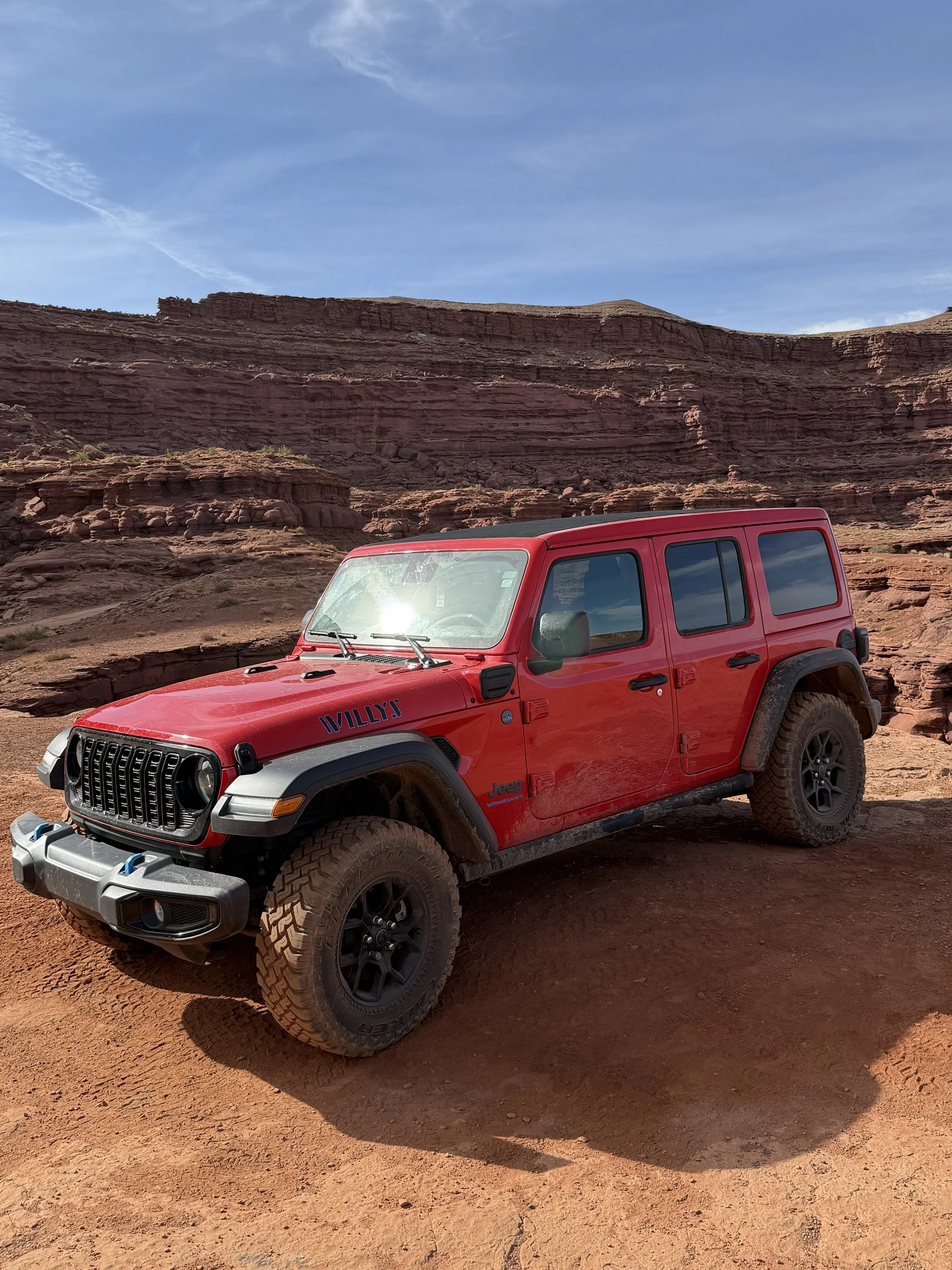 Moab Luxury Tours and Transportations' red Jeep Wrangler Willys edition parked against the dramatic sandstone canyon walls of Moab, Utah — offering guests an adventure through the region's iconic red rock backcountry.