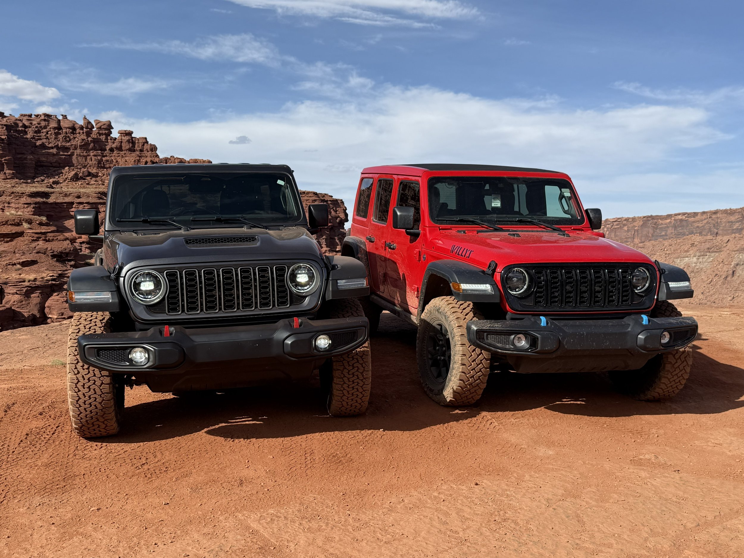 Two Jeep Wranglers, one black and one red, parked on desert terrain with rocky cliffs in the background under a partly cloudy sky.