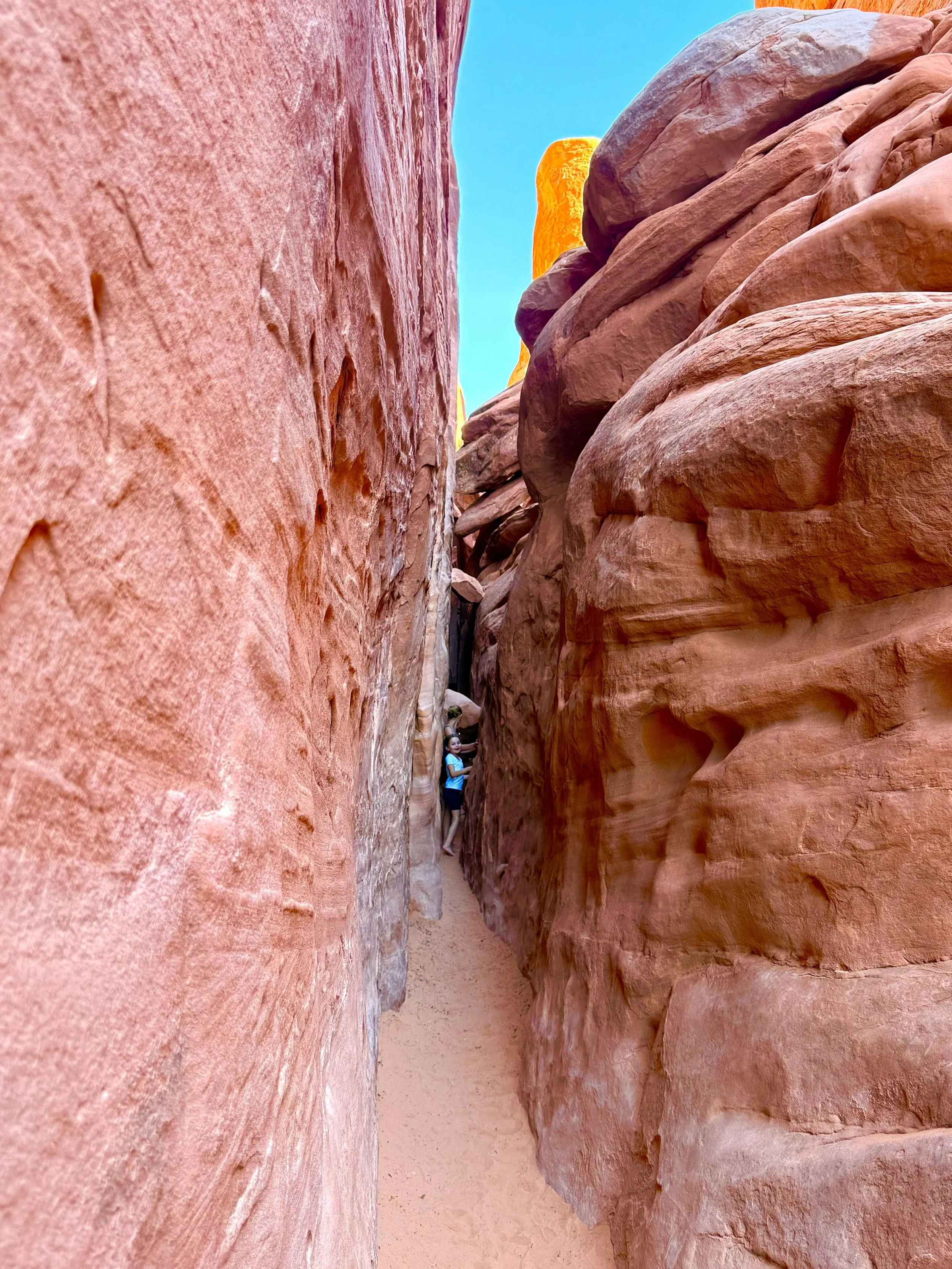 A person standing in a narrow slot canyon with tall reddish-orange rock walls on either side, wearing a blue shirt and shorts, under a bright blue sky.