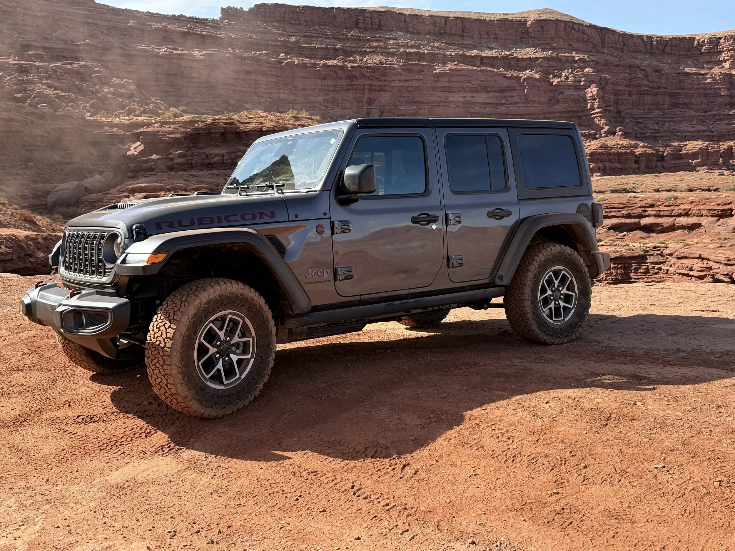 A black Jeep Rubicon SUV parked on a dirt terrain with red rock formations and cliffs in the background.