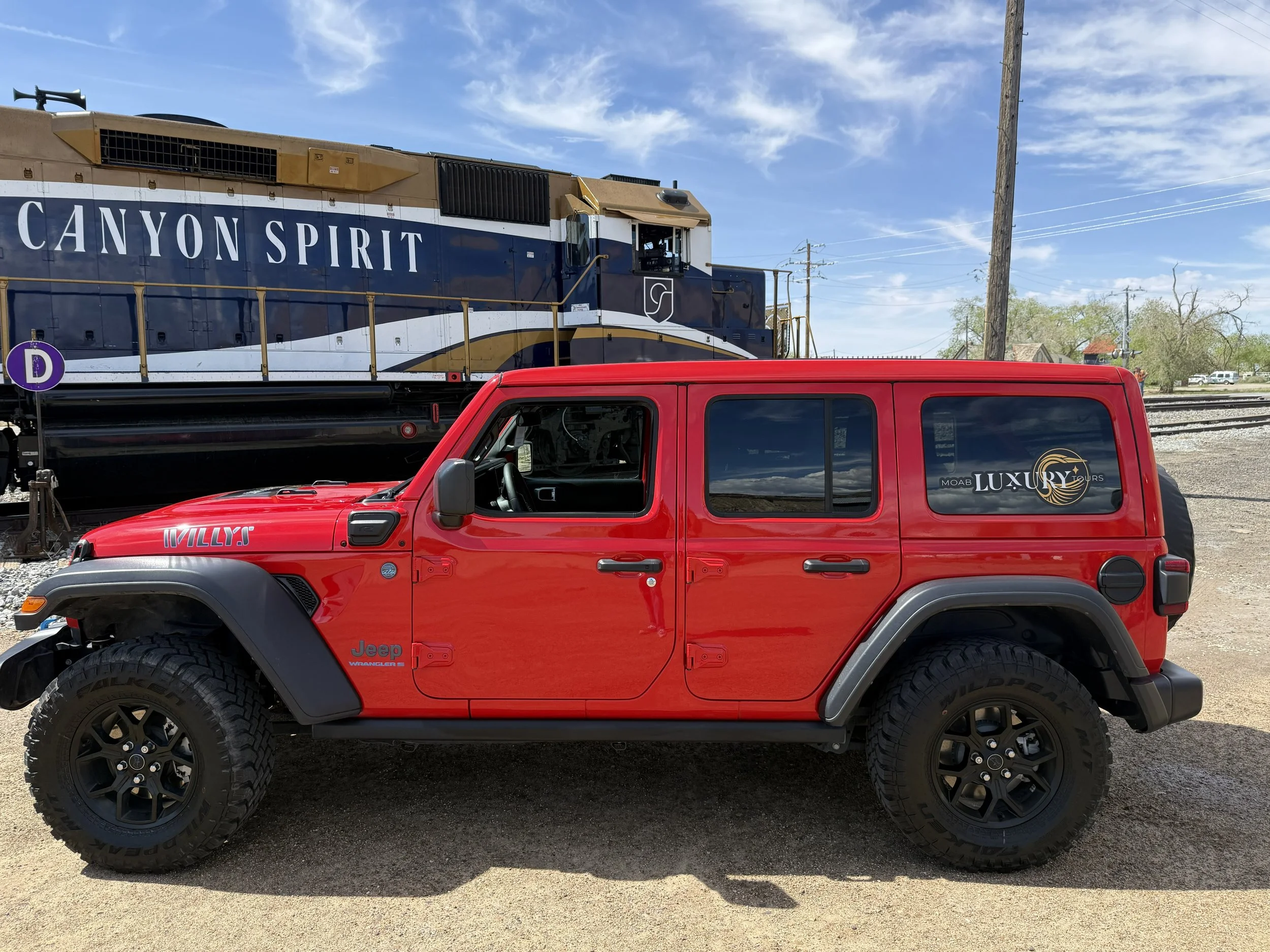 Red Jeep Wrangler parked on a dirt lot with a train in the background. The train has a blue and gold logo that reads 'Canyon Spirit' and the side door sticker says 'Luxury Tours.' The Jeep has large off-road tires and modifications, with the name 'Wi