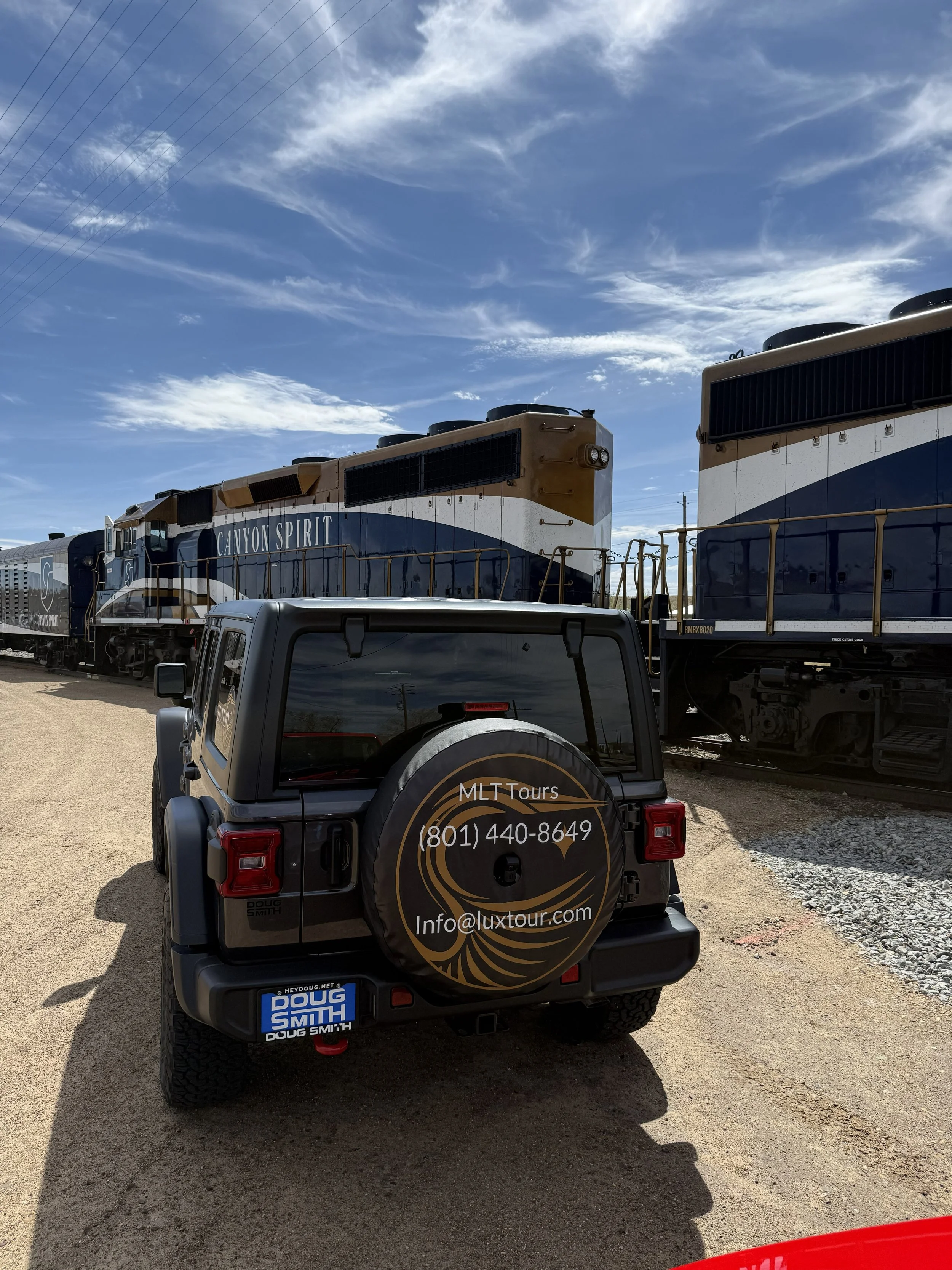 A Jeep parked on a dirt lot with a spare tire cover displaying contact information for MLT Tours, in front of a train with two diesel locomotives under a partly cloudy sky.