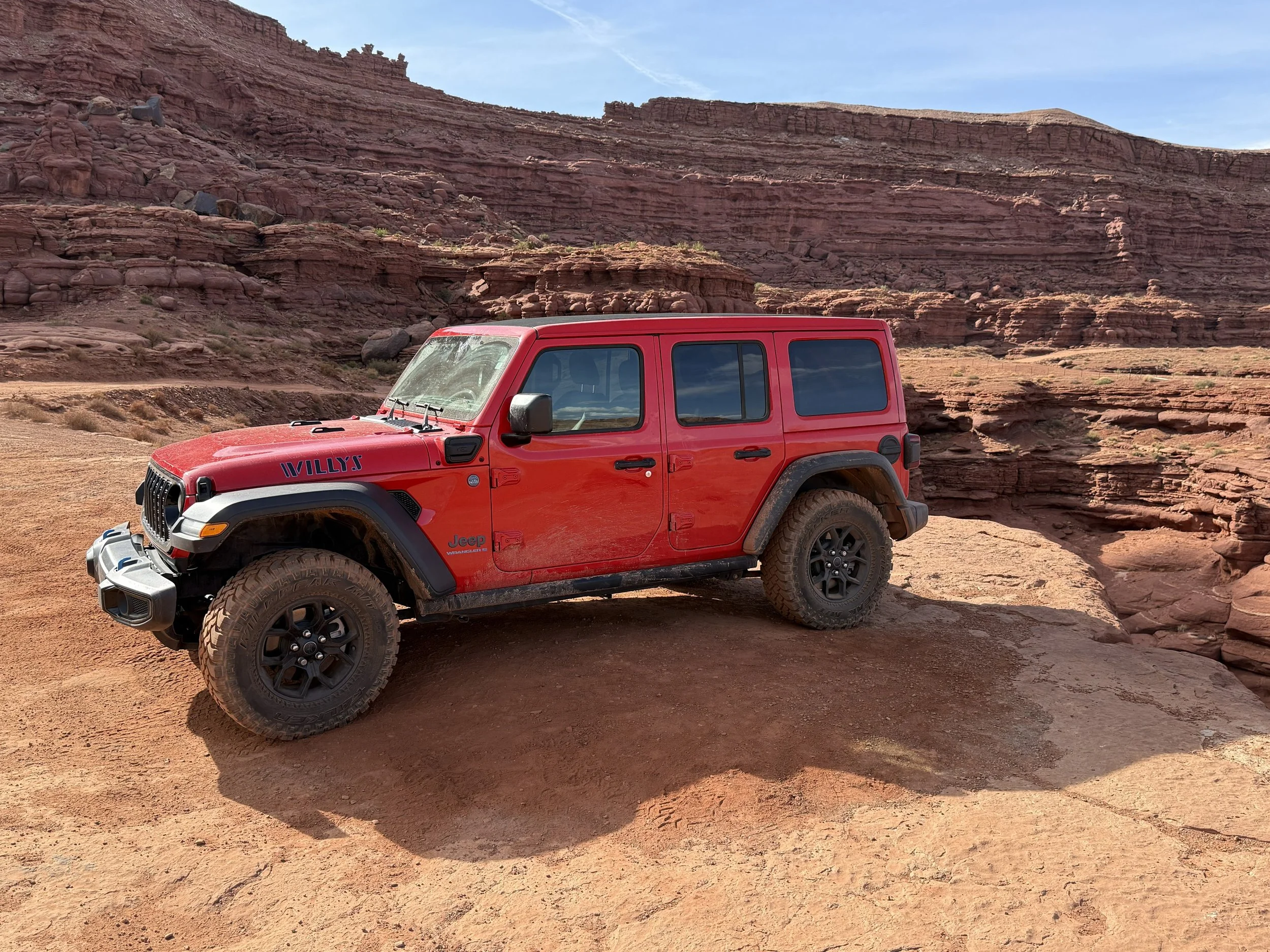 Red Jeep Wrangler Willys edition parked on dusty desert terrain with red rock formations and cliffs in the background.