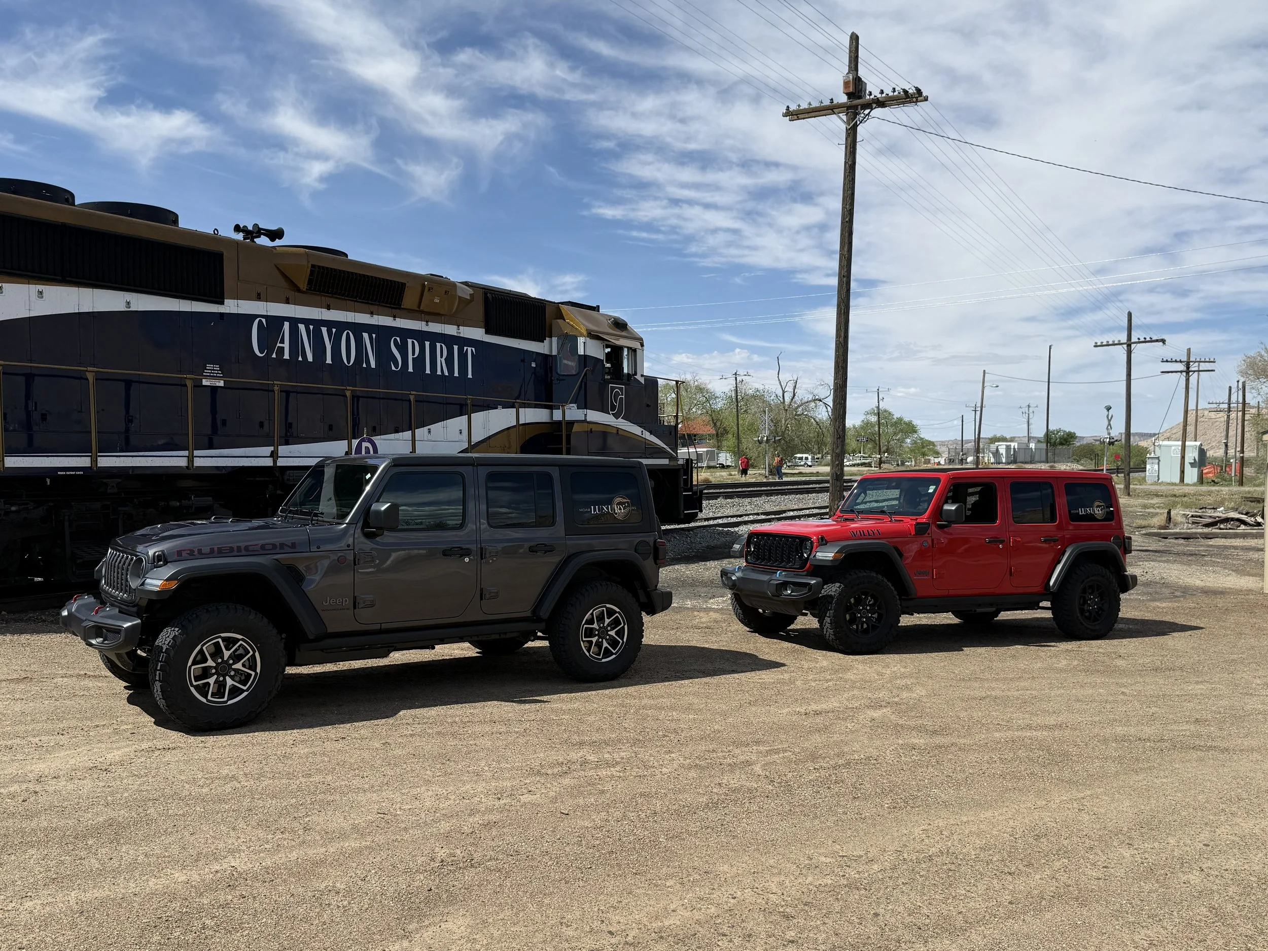 A large black and white train labeled 'Canyon Spirit' is parked on train tracks. In front of the train, there are two Jeep vehicles, one gray Rubicon and one red Willys, parked on a dirt surface. Utility poles and a few small buildings are visible in