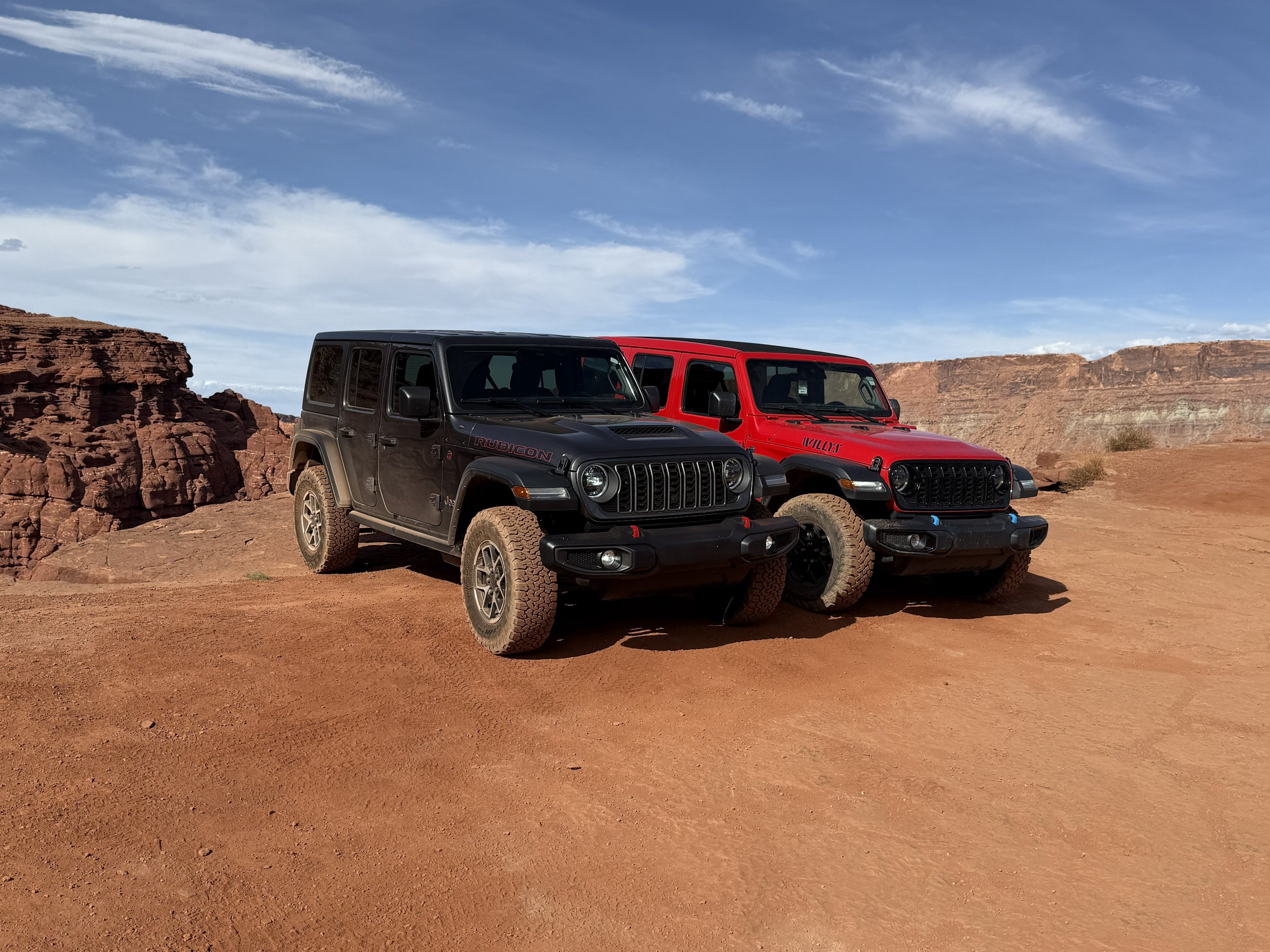 Two Jeep vehicles, black and red, parked on desert terrain with a rocky canyon background under a partly cloudy sky.