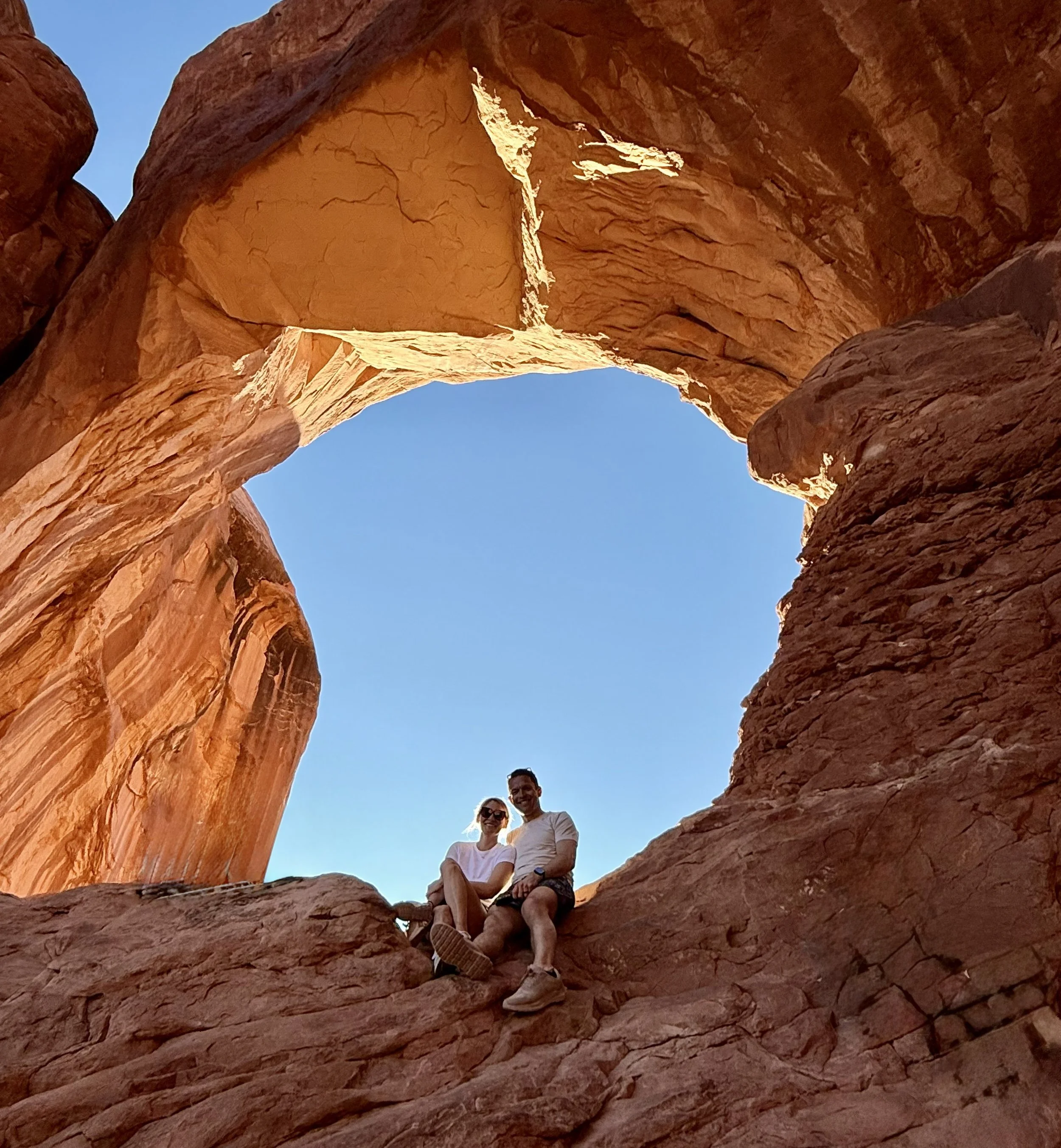 Two people sitting on red rock formations in a desert, with a natural rock arch overhead and a clear blue sky.