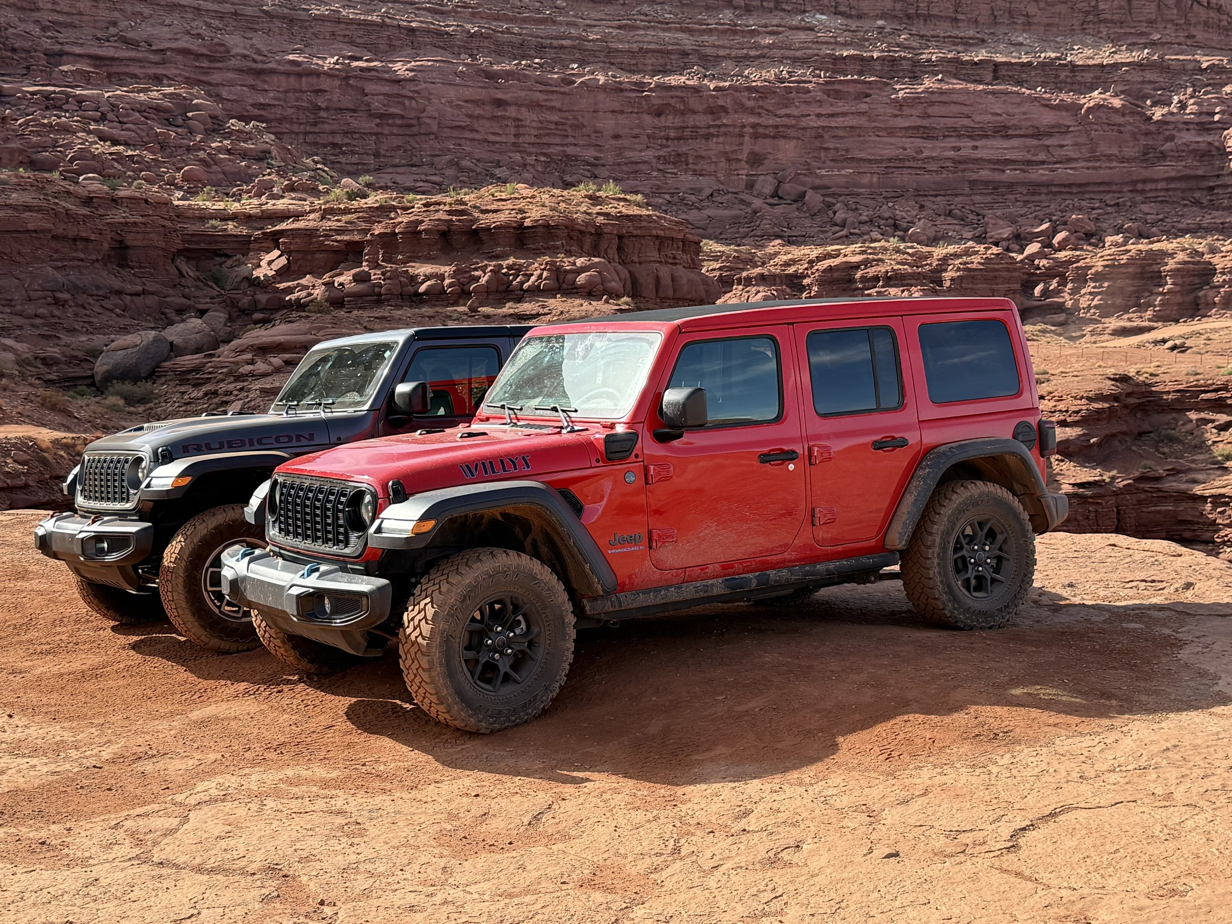 Two Jeep vehicles parked on a dirt trail with red rock formations in the background.