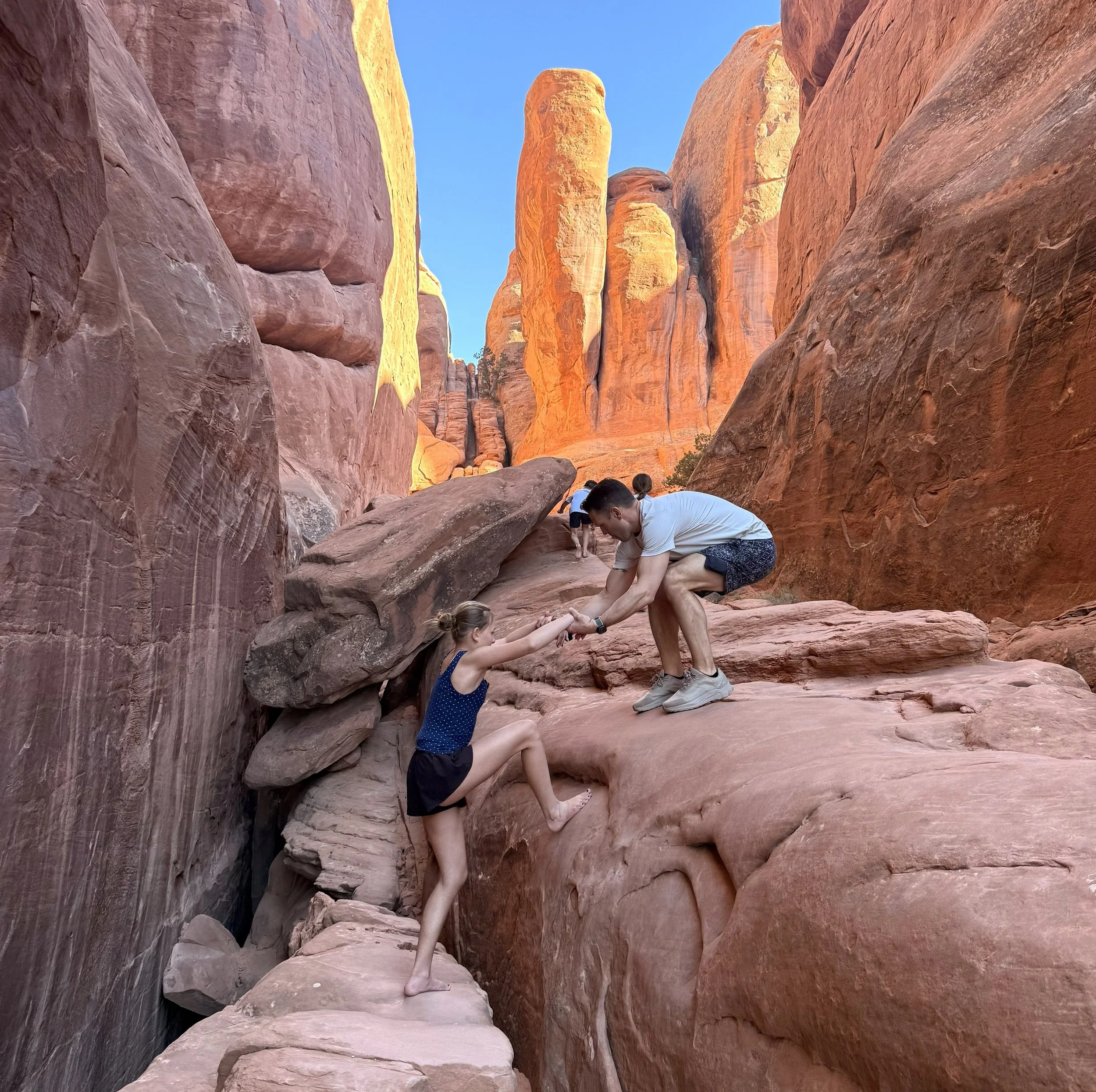 A man helping a young girl climb a rock in a canyon with tall red rock formations.