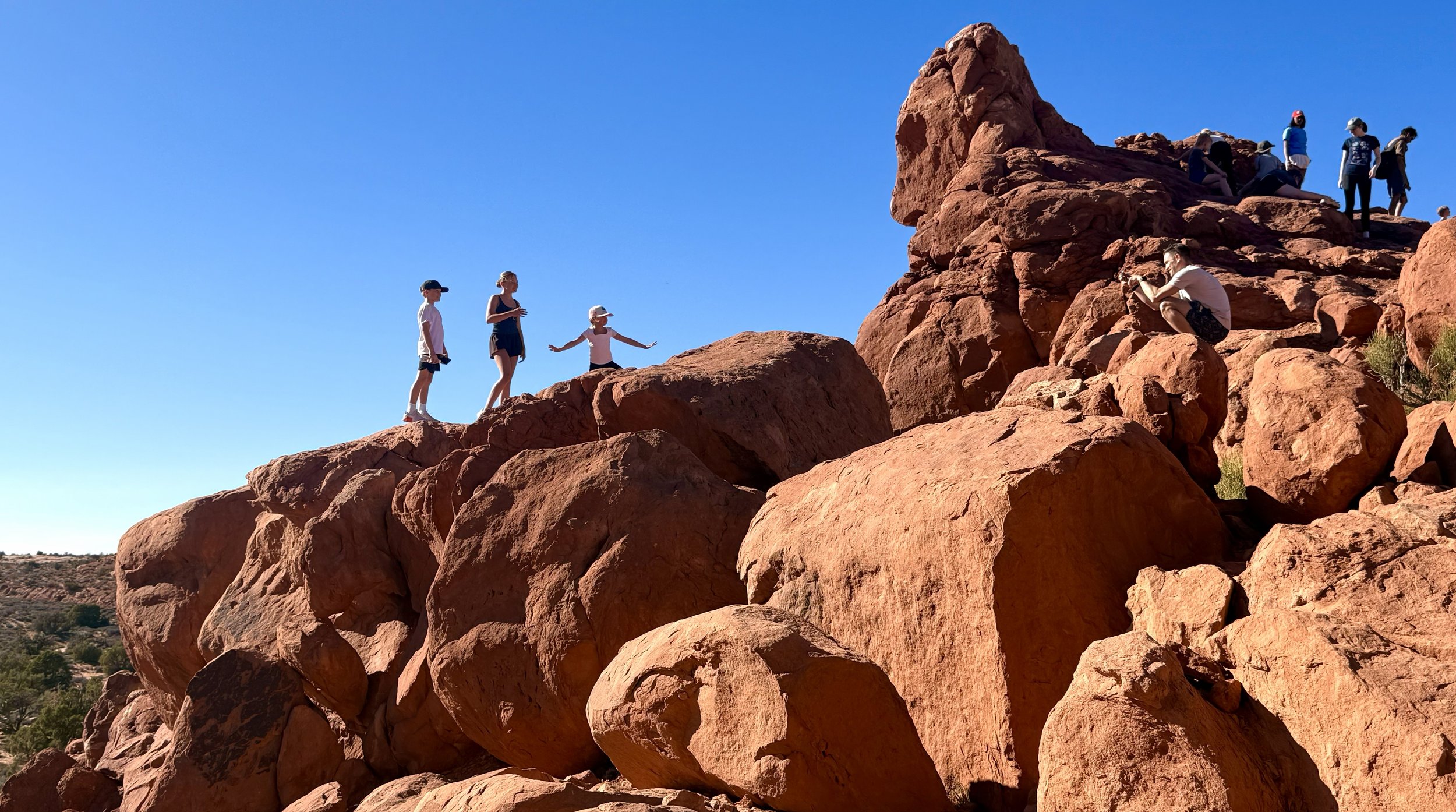 People climbing and sitting on red rocks in a desert landscape under a clear blue sky.