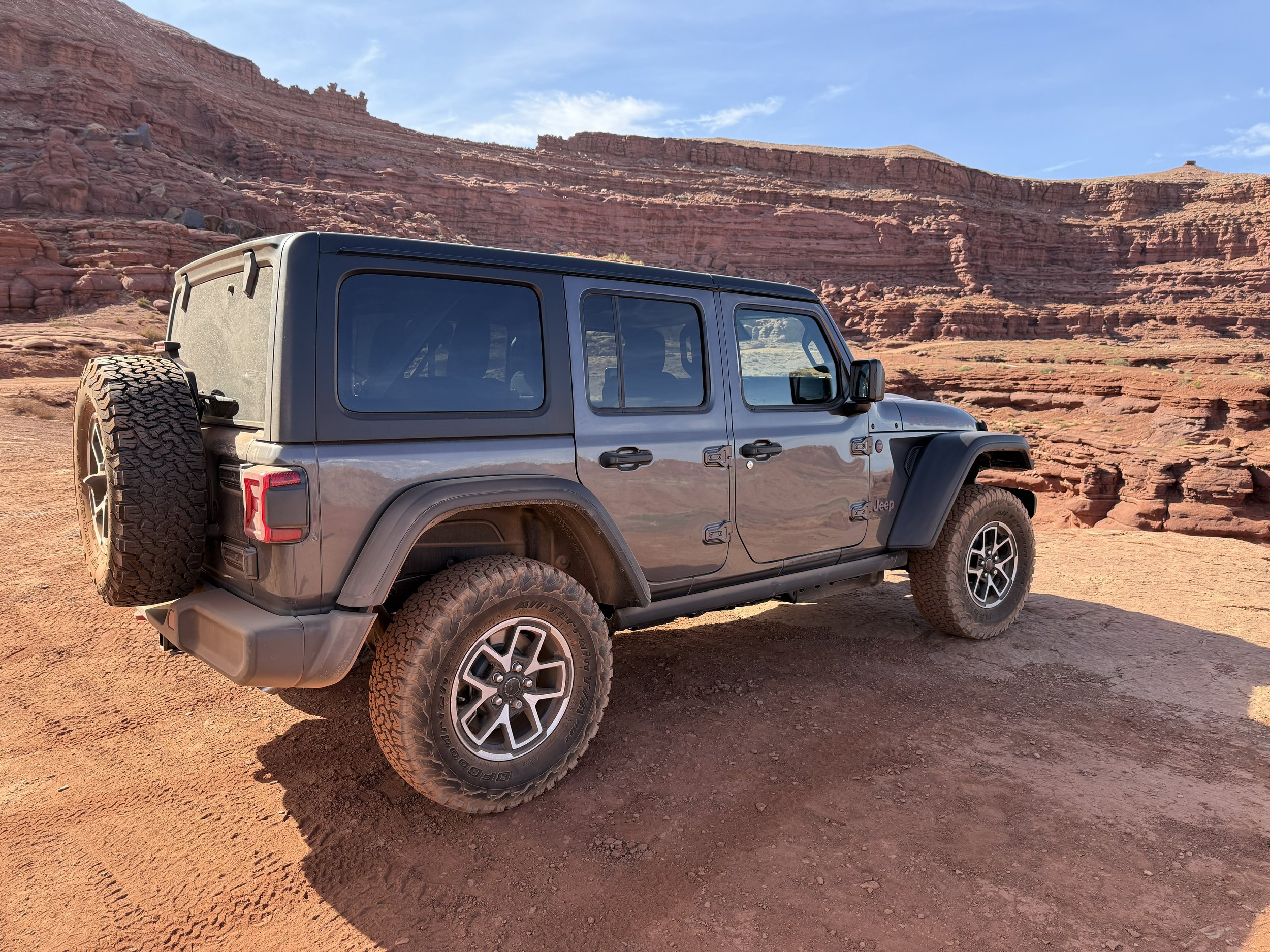 A gray Jeep Wrangler parked on a dirt surface in a desert landscape with red rock formations and blue sky.