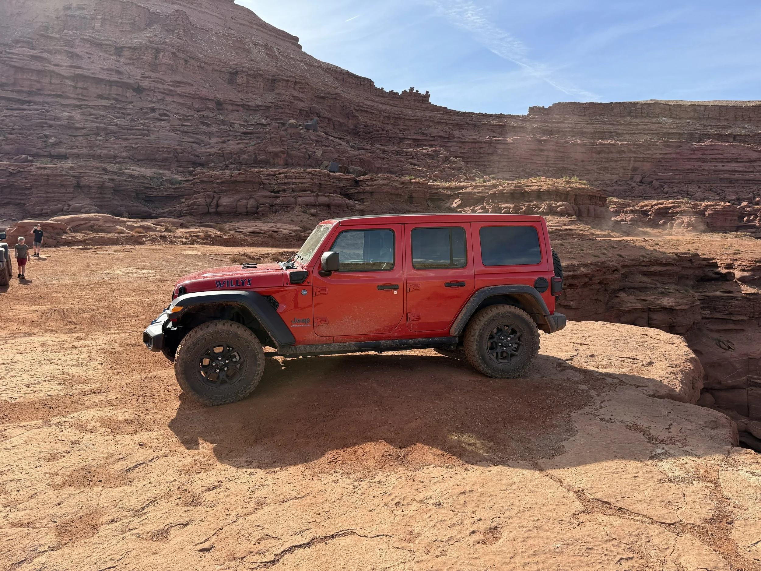 A red Jeep Wranger with black wheels parked on a rocky desert landscape with cliffs in the background and a few people in the distance.