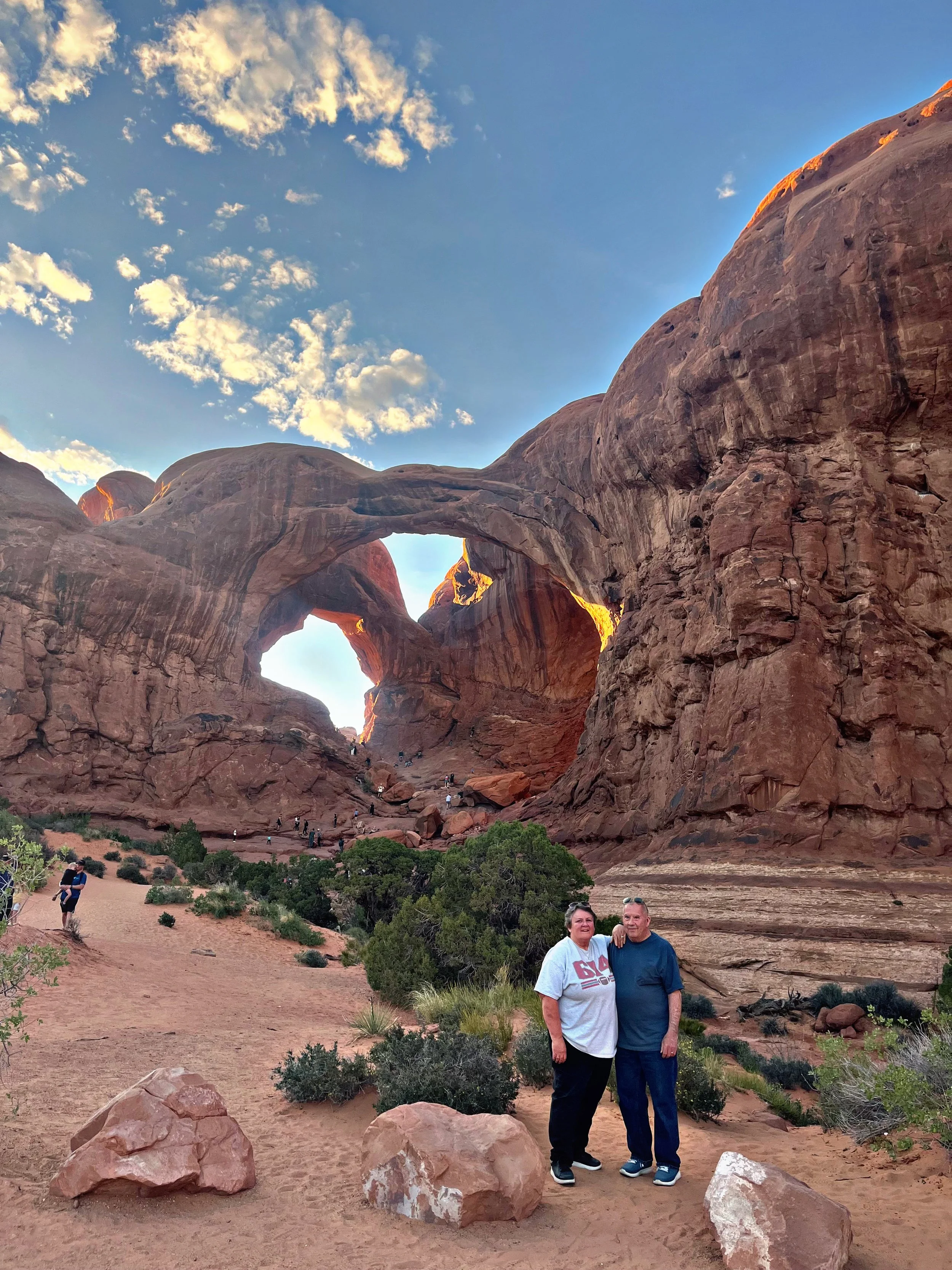 A couple standing close together in front of the Arches National Park's famous rock formations with a large natural stone arch behind them, under a partly cloudy sky.