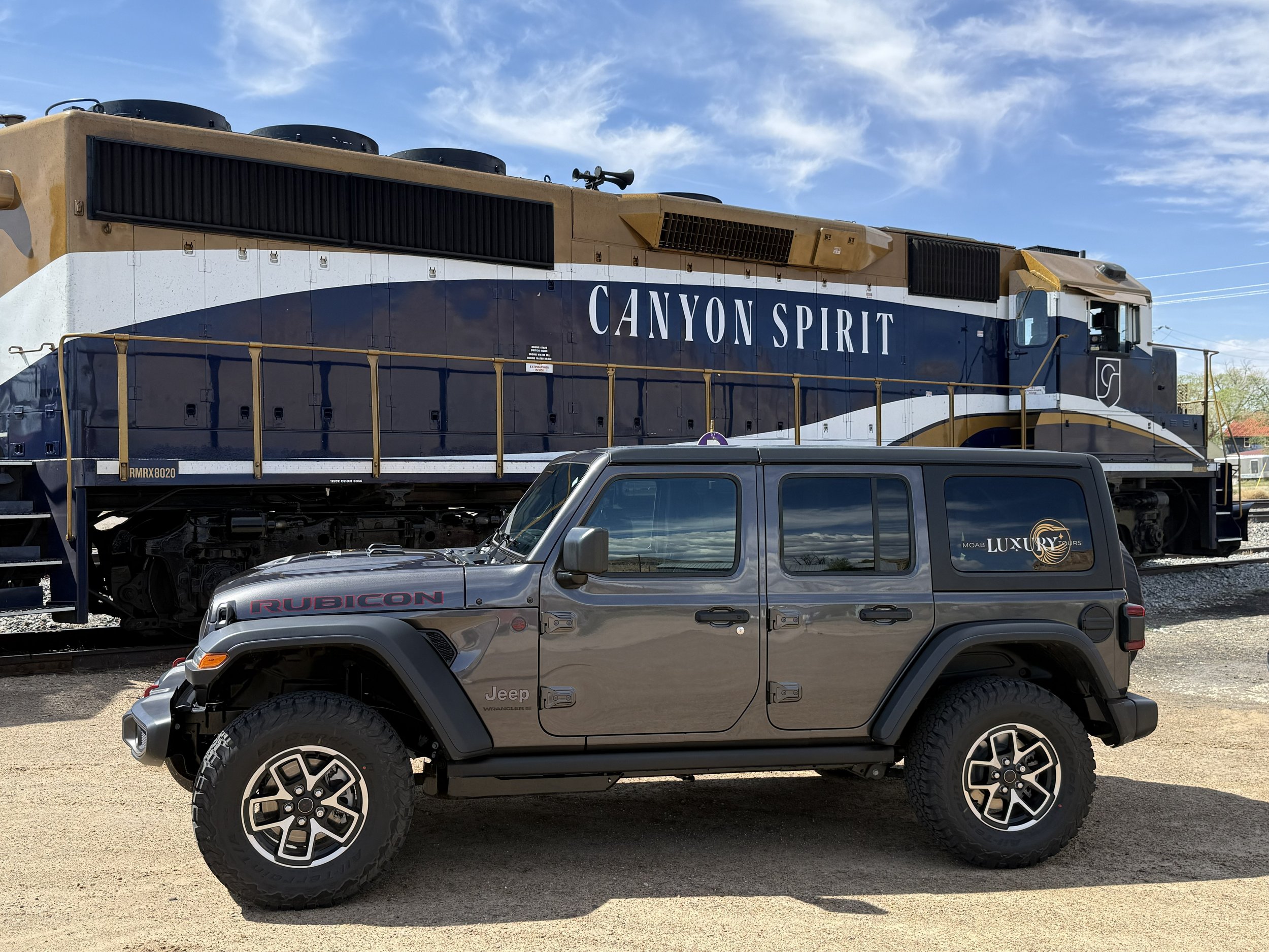 A gray Jeep Rubicon SUV with all-terrain tires parked in front of a freight train engine painted in blue and white, with "Canyon Spirit" written on its side, on a sunny day with a blue sky.