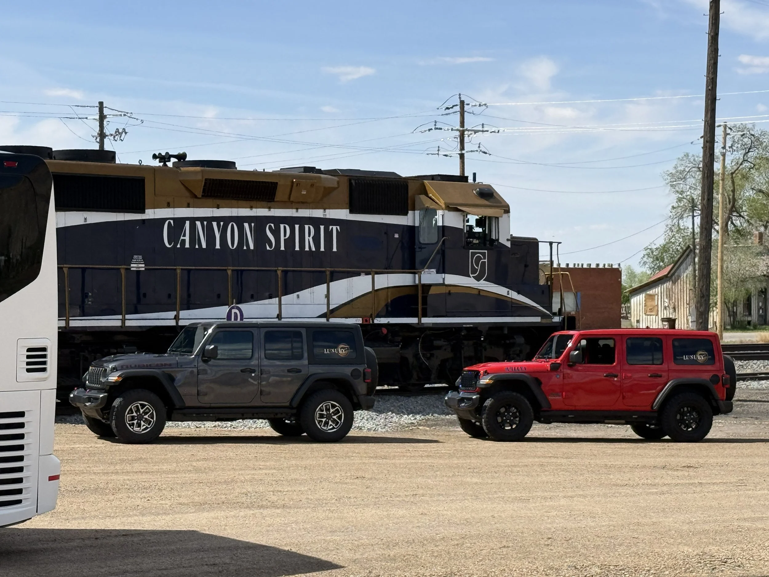 A moving train with the words 'Canyon Spirit' on its side, two parked SUVs (one black, one red) in front of the train on a gravel area, utility poles, and small buildings in the background under a partly cloudy sky.