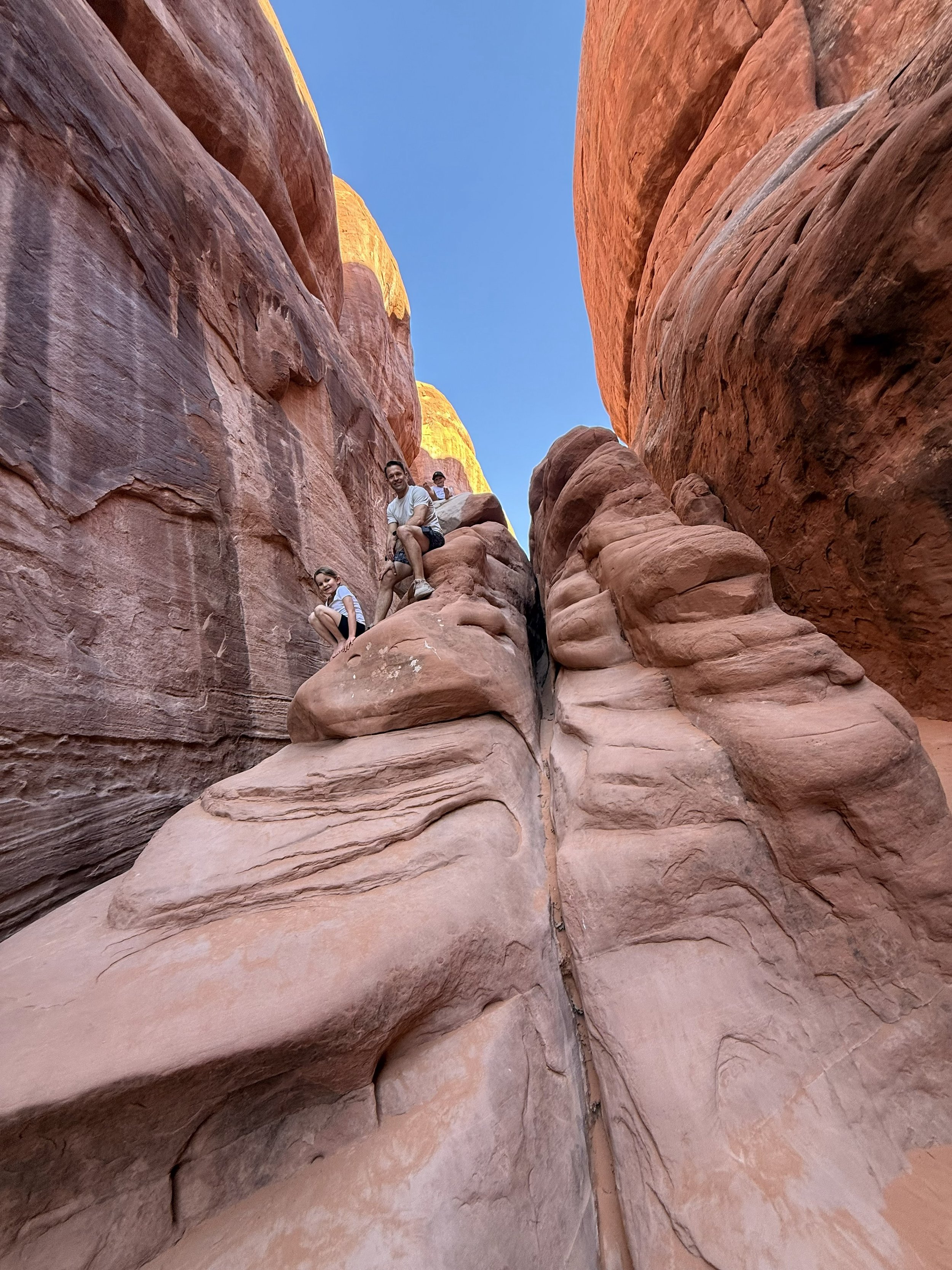 Three children climbing on sandstone rocks in a narrow canyon with red and pink hues, blue sky overhead.