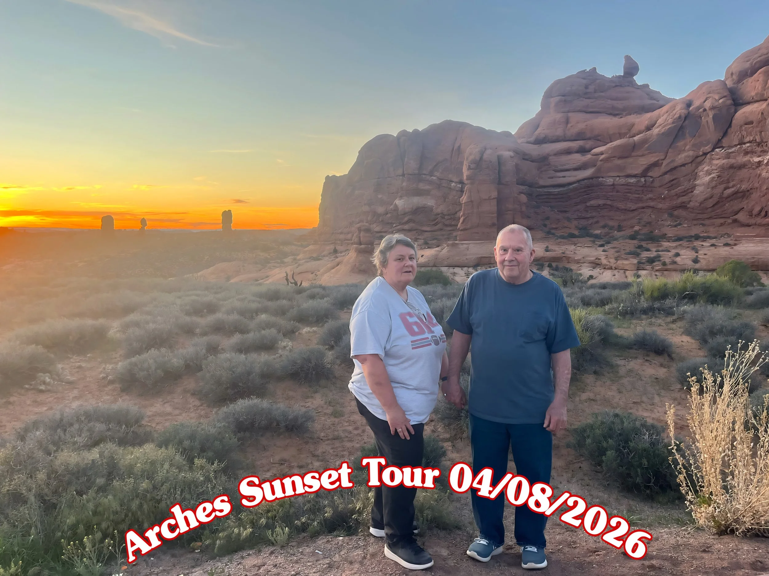 A couple standing in a desert landscape at sunset with large red rock formations and sparse vegetation, with the text 'Arches Sunset Tour 04/08/2026' overlaid at the bottom.