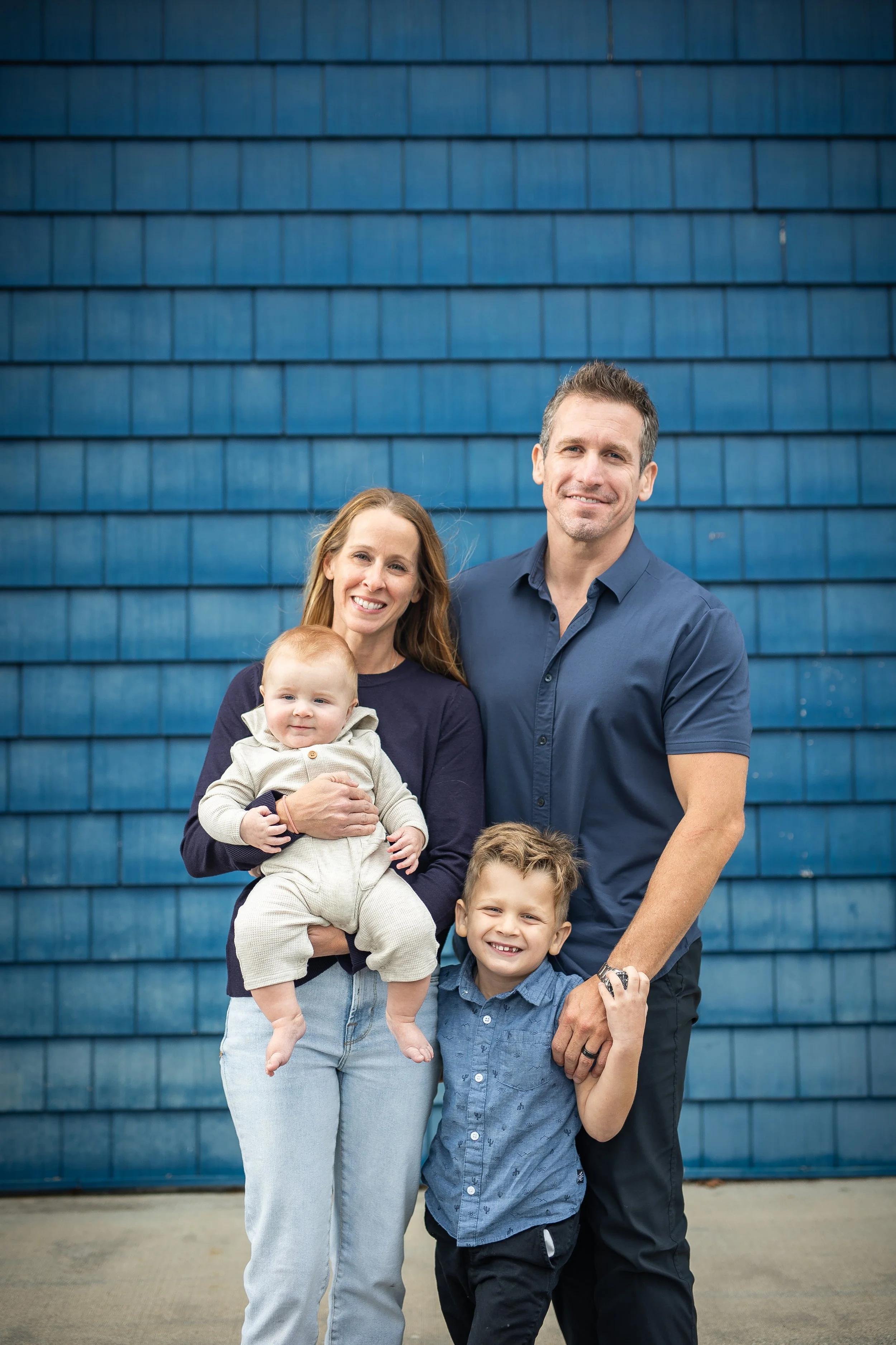 Family of four standing outdoors in front of a blue building. The mother is holding a baby, and the father and older son are standing next to them, all smiling.