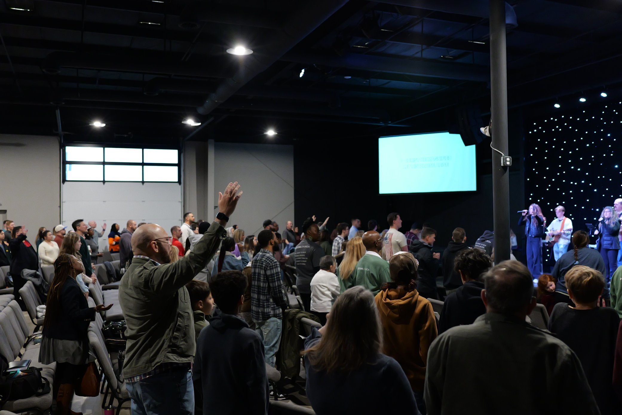 People gathered in a large indoor space, some raising their hands, attending a stage event with a band performing and a bright screen on stage.