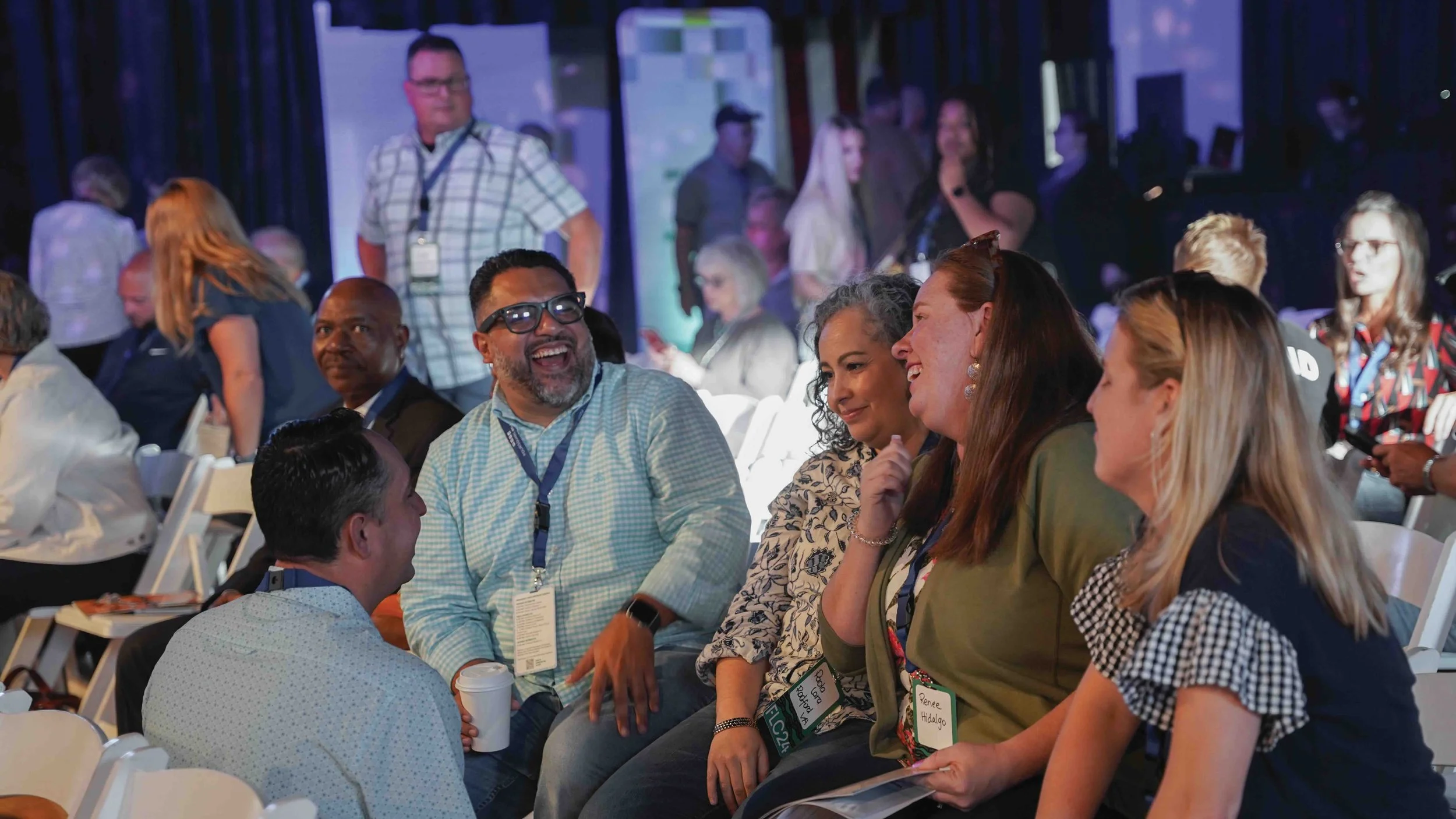 A diverse group of people seated in a conference, engaging in conversation and laughter, with some wearing conference badges and a large screen or projection in the background.