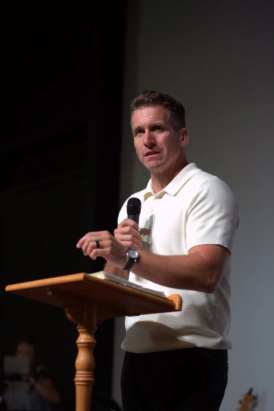 A man in a white polo shirt is speaking into a microphone while standing at a wooden podium, with a notebook on top, in a dimly lit room.