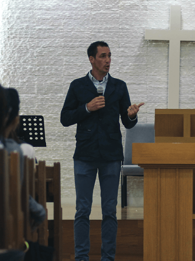 A man speaking into a microphone in a church with a cross on the wall behind him.