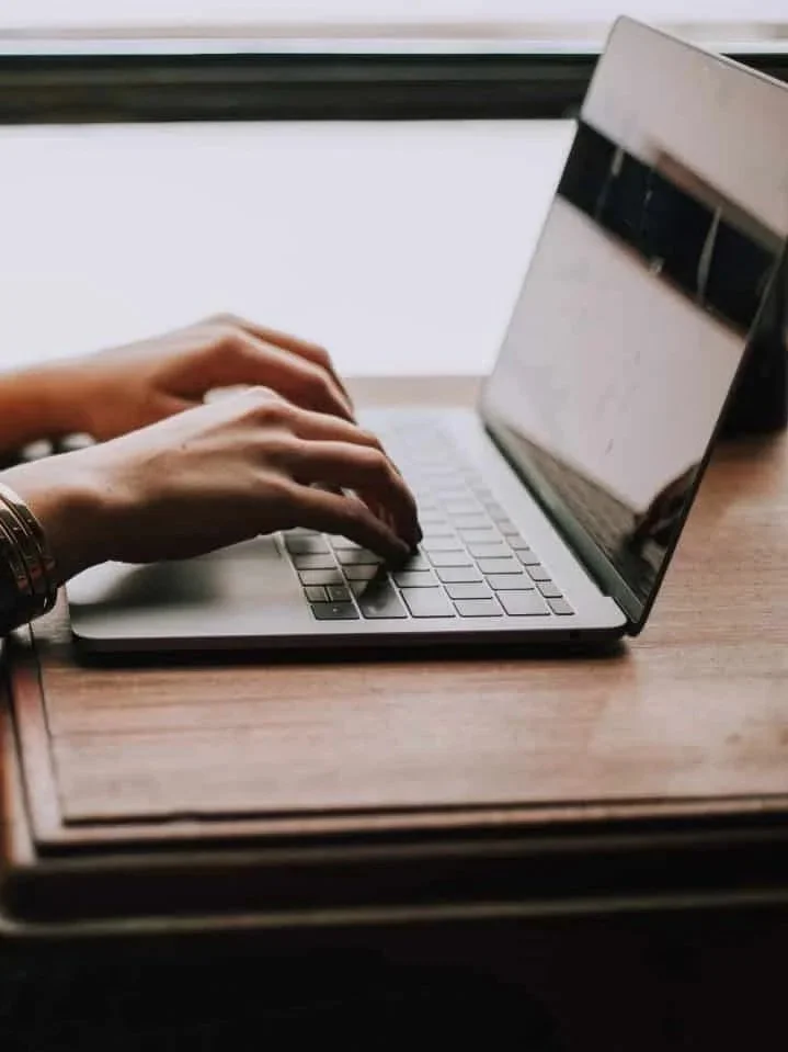 Person typing on a laptop keyboard at a wooden desk near a window.
