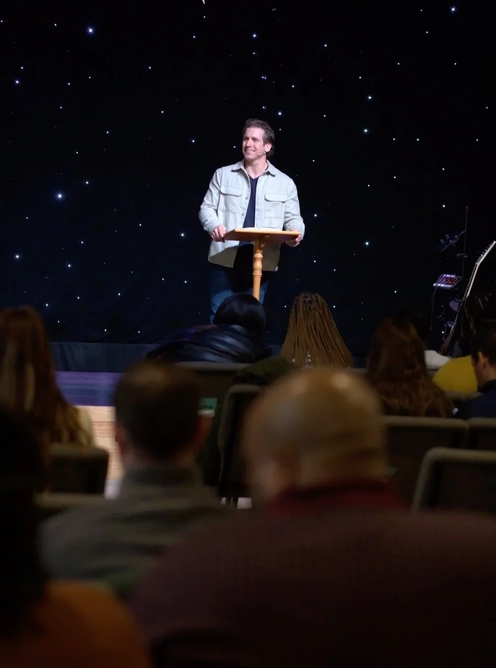 A man standing behind a lectern on a stage with a starry backdrop, giving a presentation or speech to an audience.