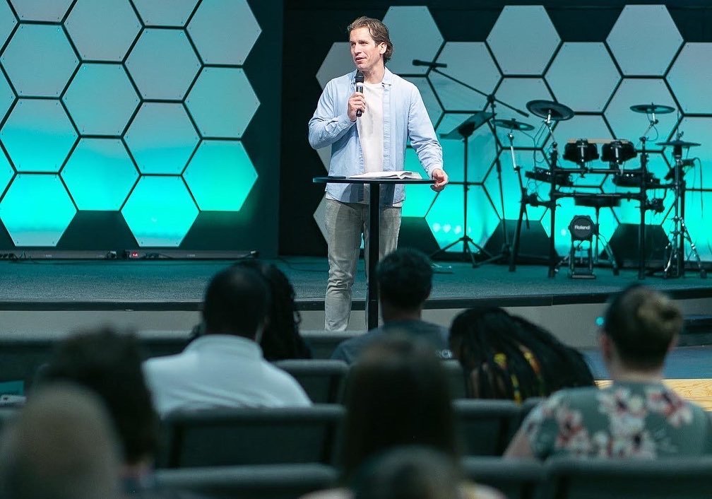 A man in casual clothing speaking at a podium with a microphone to an audience in a church or conference setting, with a backdrop of hexagonal shapes and musical instruments.
