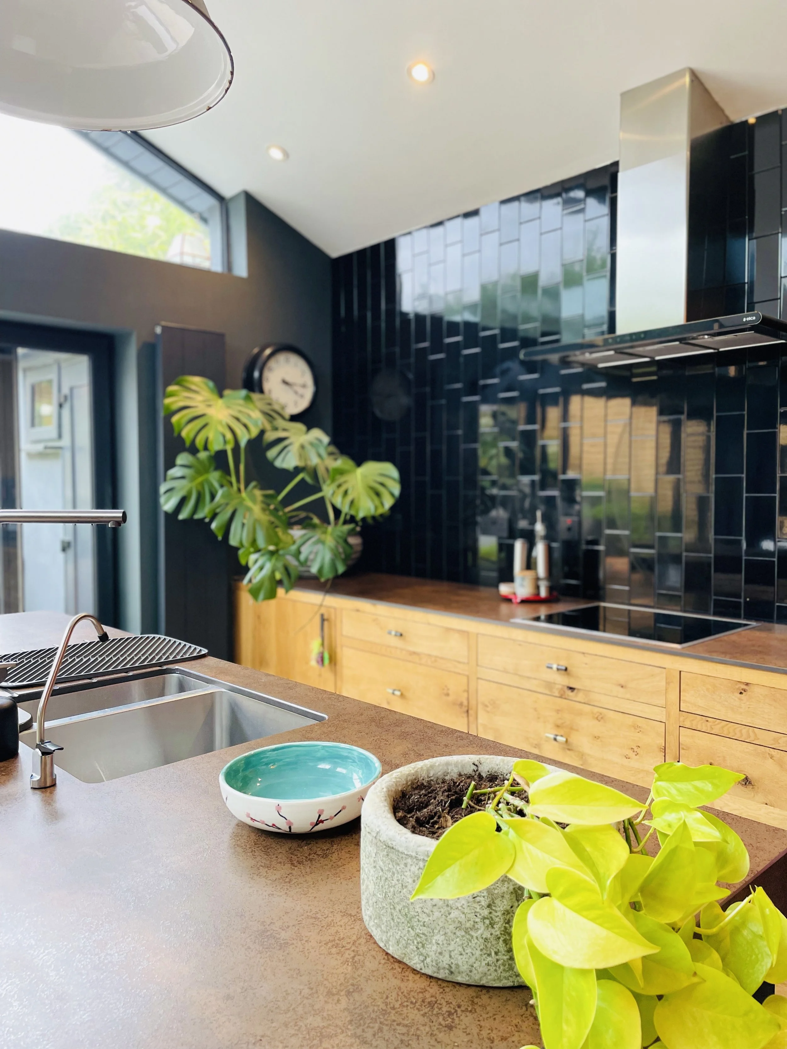 Modern kitchen with black tiled backsplash, wooden cabinets, a silver range hood, green houseplants, and a beige countertop.