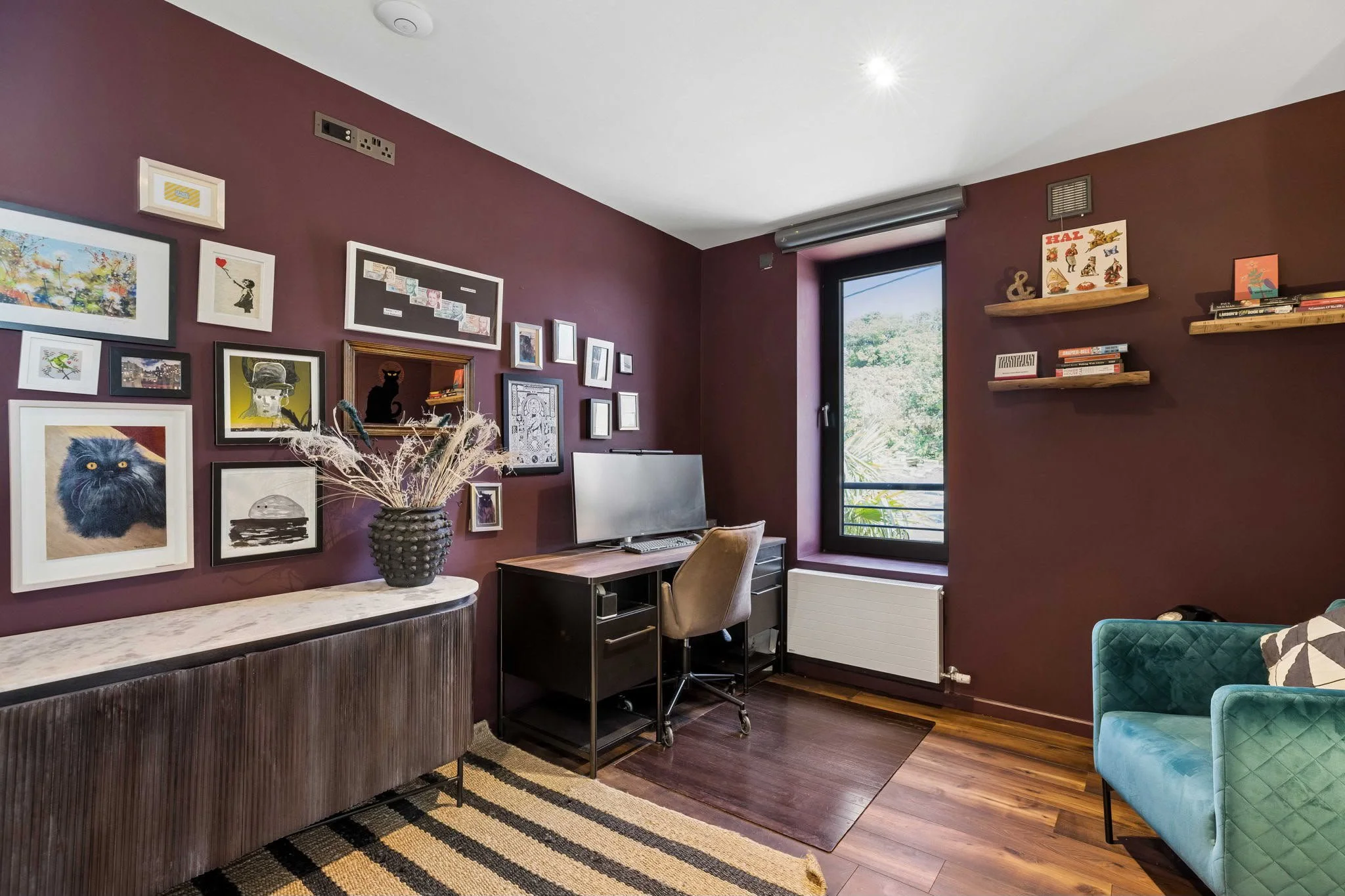 Interior of a cozy living or office space with dark purple walls, decorated with various framed artwork, photos, and decorative items. There is a desk with a monitor and chair, a window letting in natural light, and floating shelves with books and de