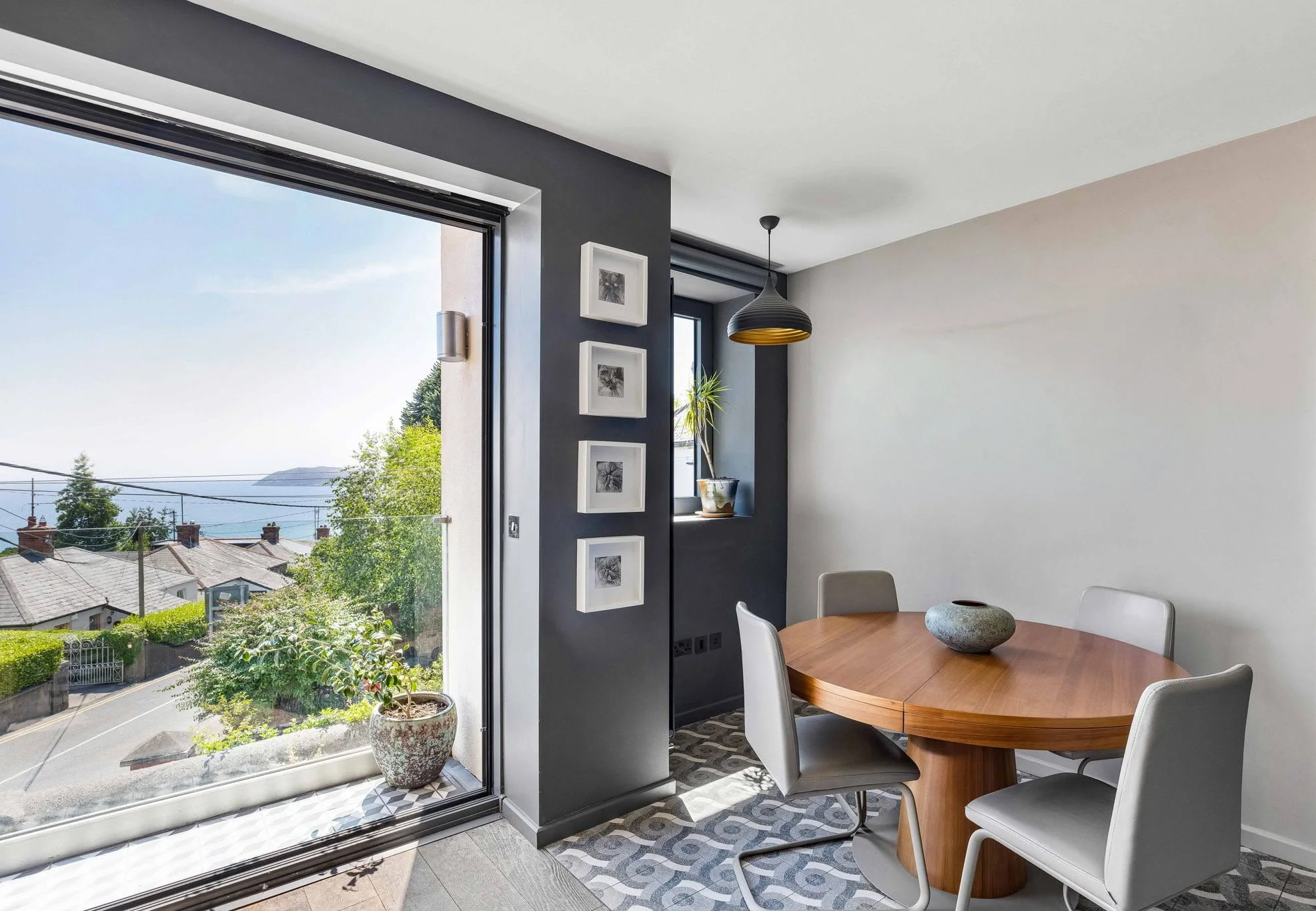 View from inside a modern dining area with large sliding glass door opening to a balcony and scenic neighborhood view. The room features a round wooden table with four white chairs, a decorative stone bowl, framed artwork on a dark accent wall, a pen