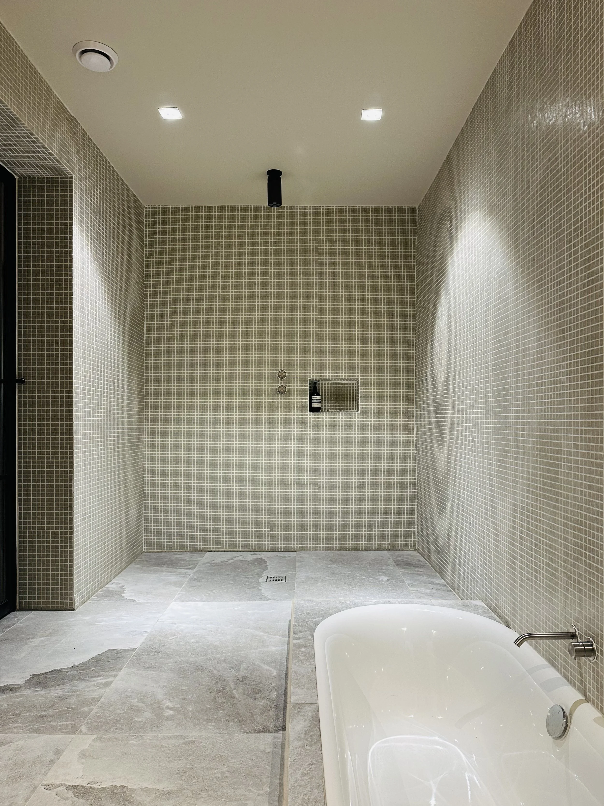 Modern bathroom with a corner shower area with beige small mosaic tiles on the walls, a small recessed shelf with soap, a ceiling light, and a bathtub with a faucet in the foreground.