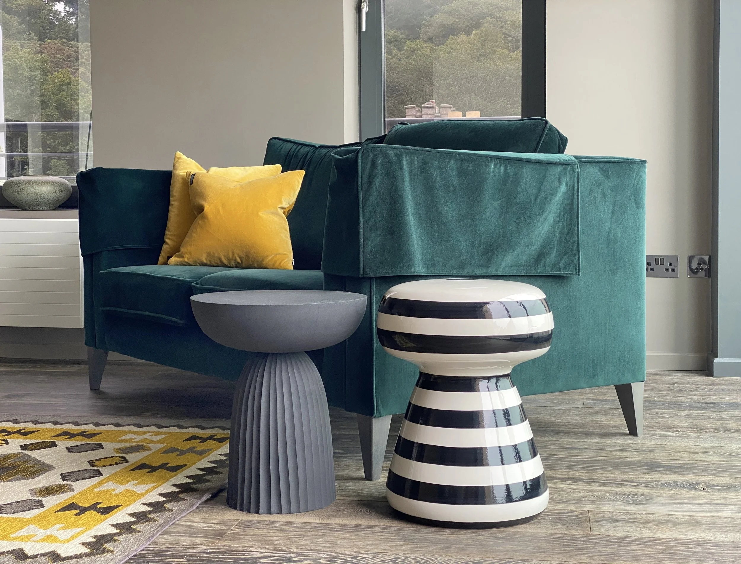 A modern living room with a teal velvet sofa, yellow and navy pillows, a round gray ottoman, and a black and white striped side table. There's a large window with a view of trees and a patterned yellow and gray rug on the wooden floor.